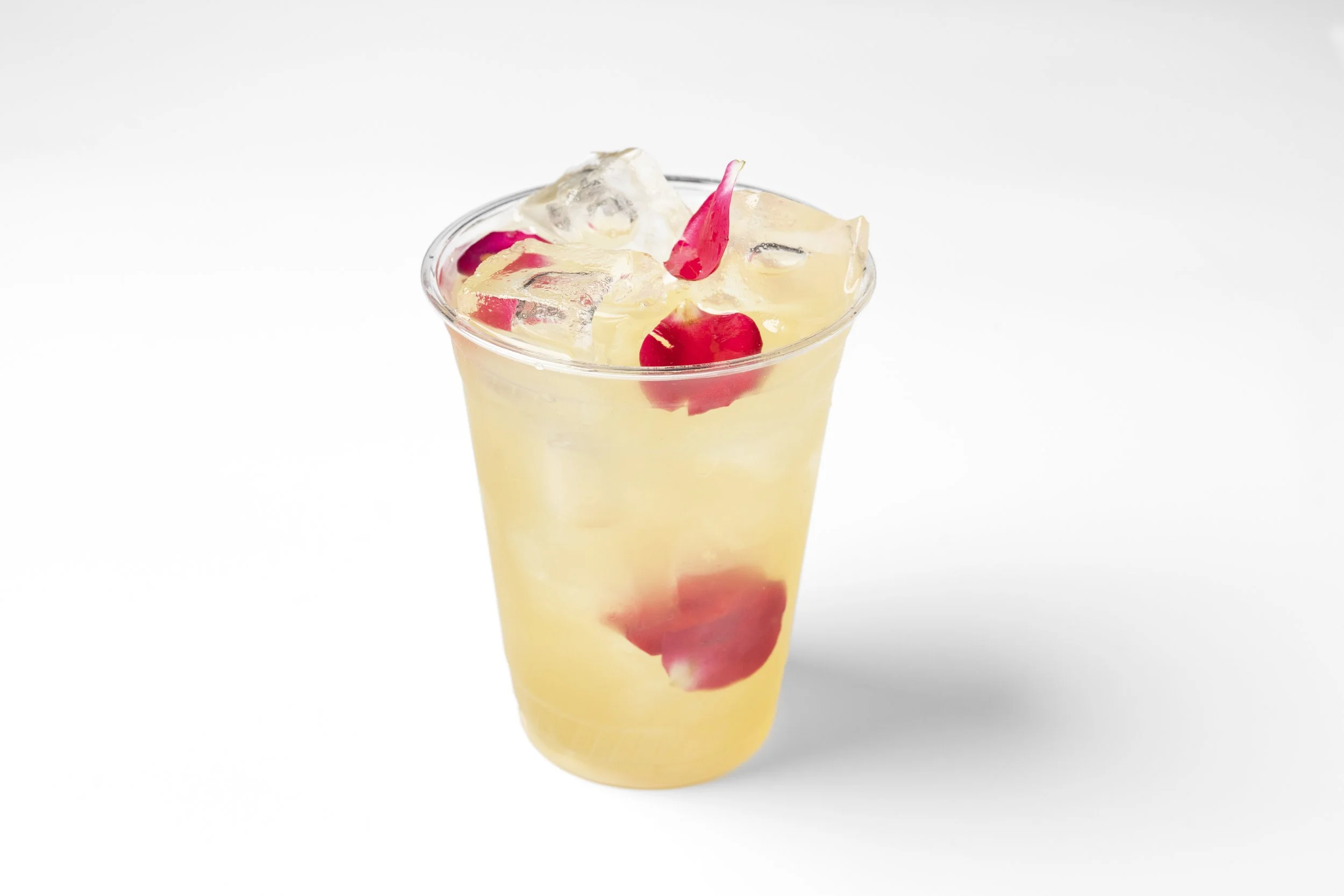 A clear plastic cup filled with a yellow beverage, ice cubes, and red rose petals