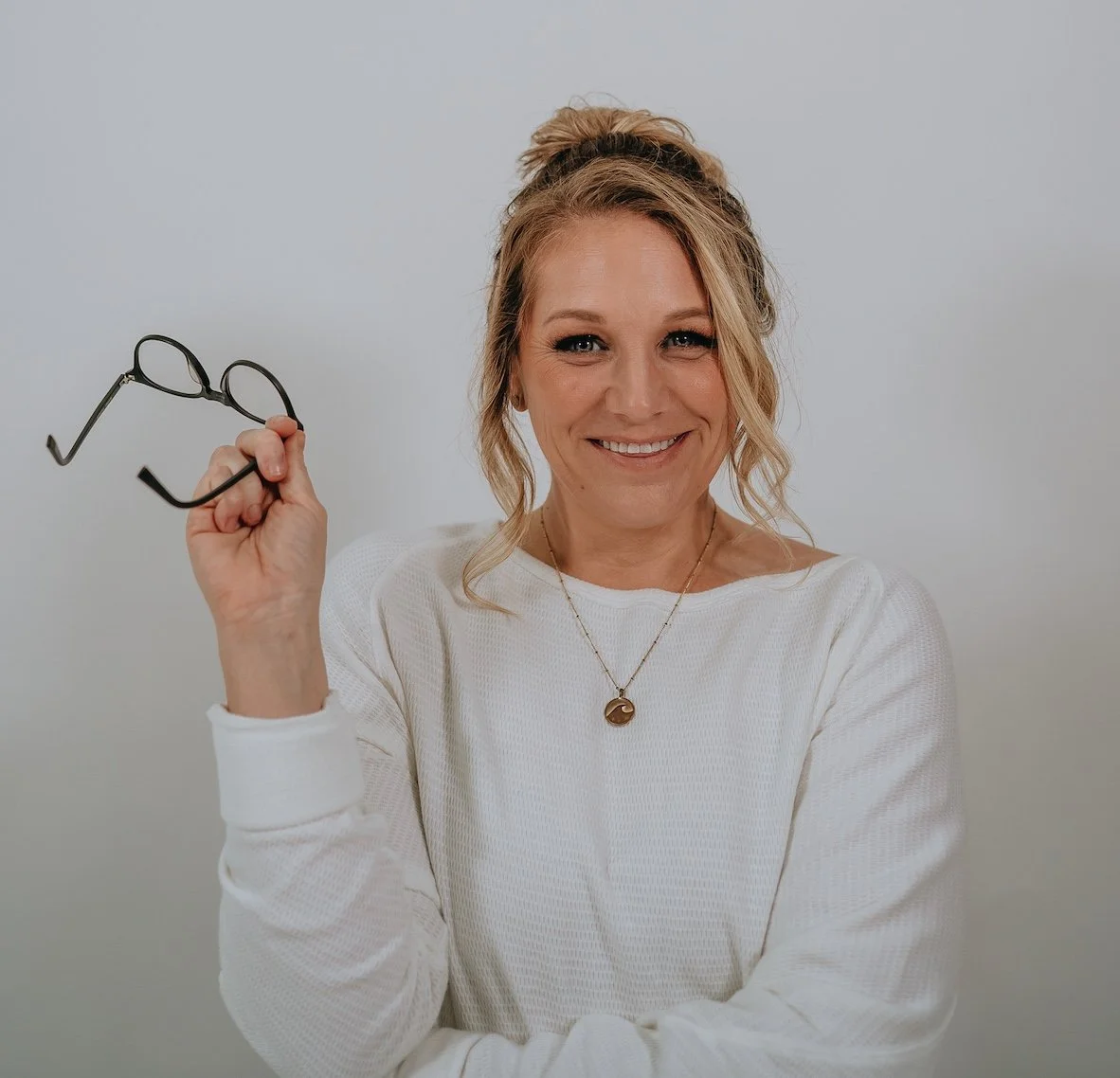 A woman with blonde hair tied in a bun, smiling, holding black-rimmed glasses in her right hand, wearing a white sweater and a necklace with a wave pendant, standing against a plain gray background.
