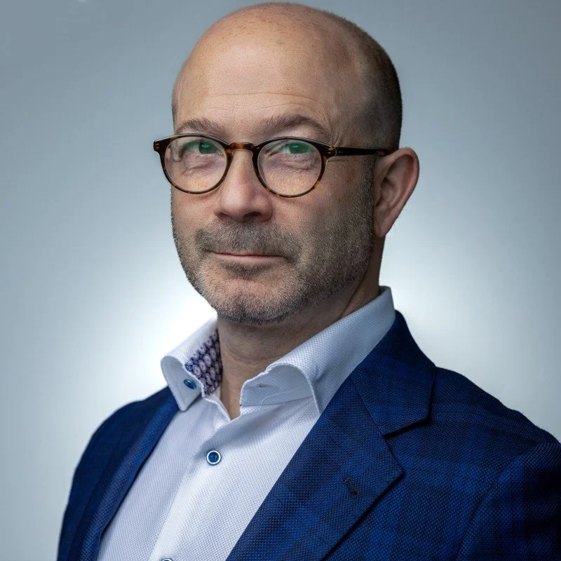A middle-aged man with glasses, wearing a white dress shirt and a dark blue blazer, looking at the camera against a plain gray background.