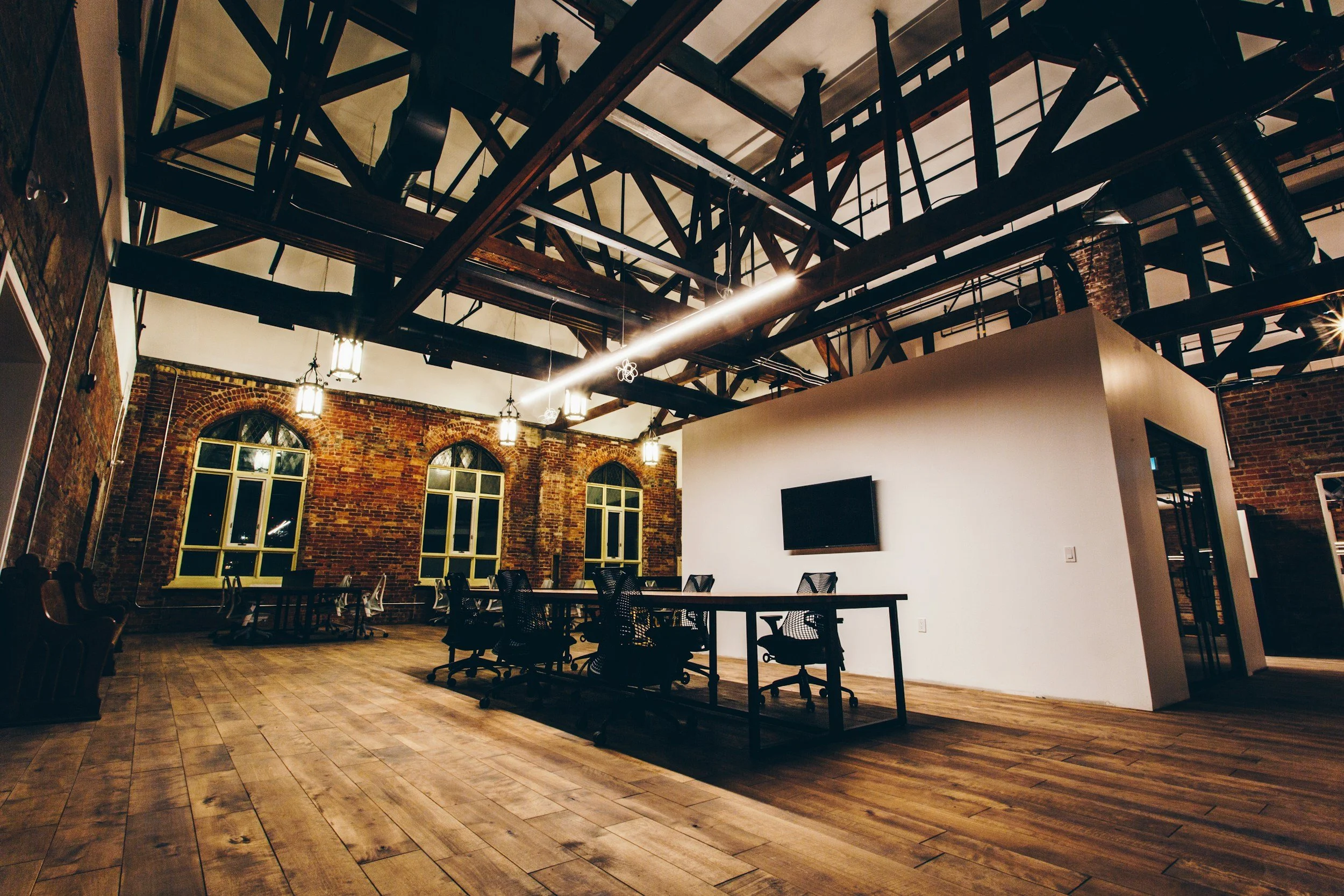 Interior of a modern office with brick walls, large windows, wooden floors, a white partition wall with a mounted television, and black chairs around tables, featuring exposed ceiling beams and industrial-style lighting.