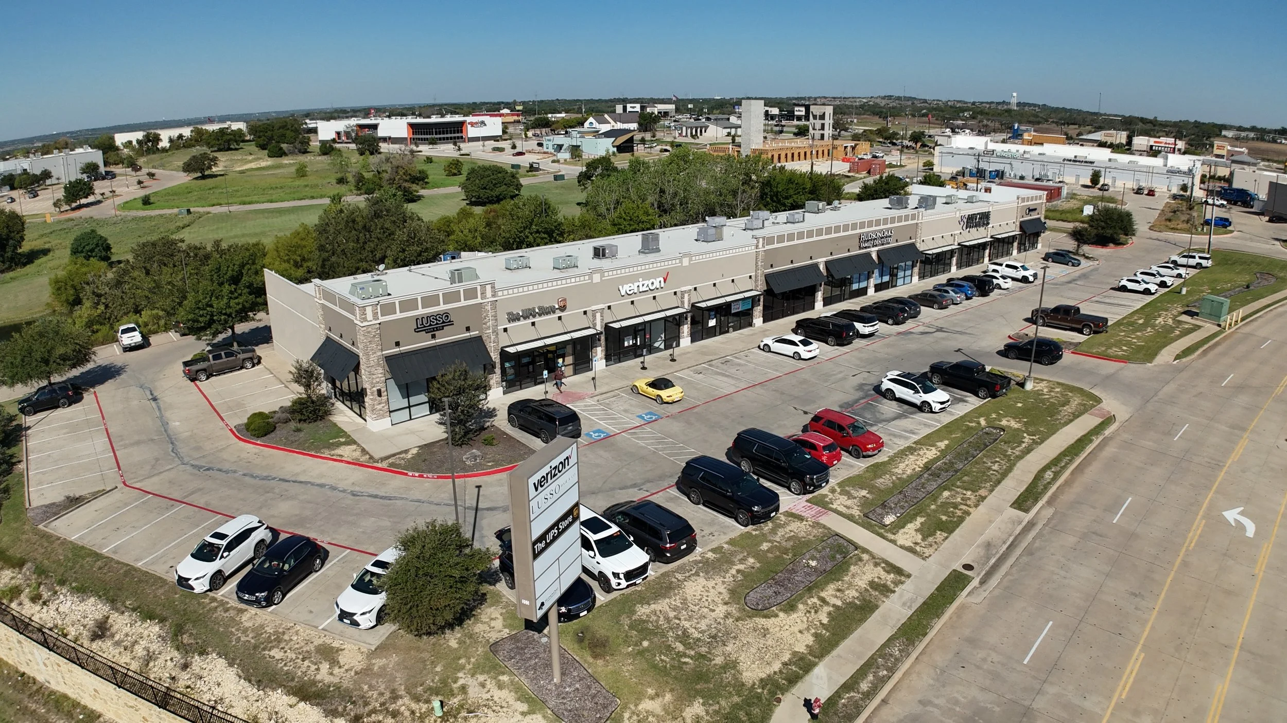 An aerial view of a shopping plaza with parked cars, trees, and surrounding buildings.