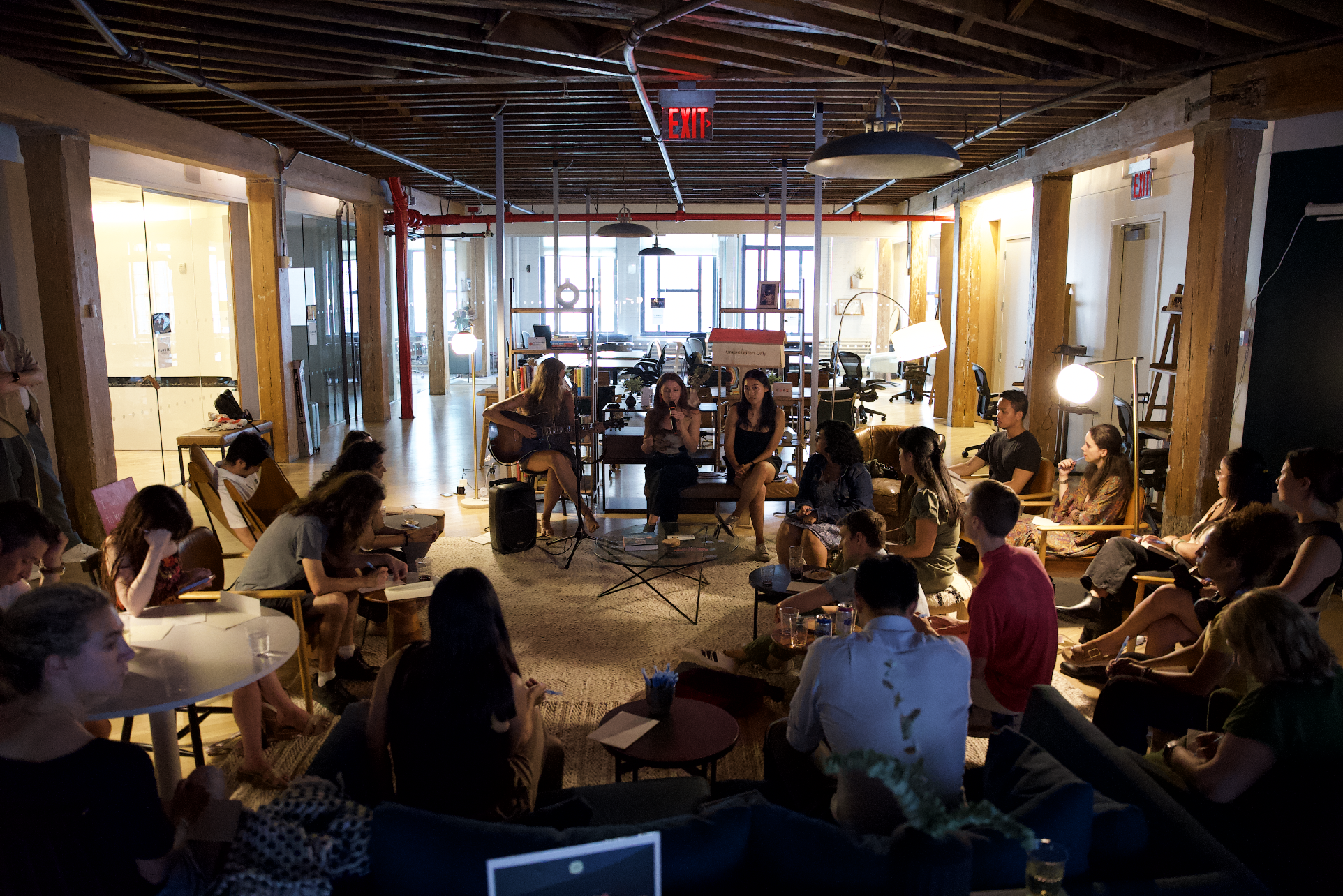 A group of people gathered in a cozy indoor space for a live performance, with some sitting on chairs and couches, and a musician playing guitar and singing on stage.