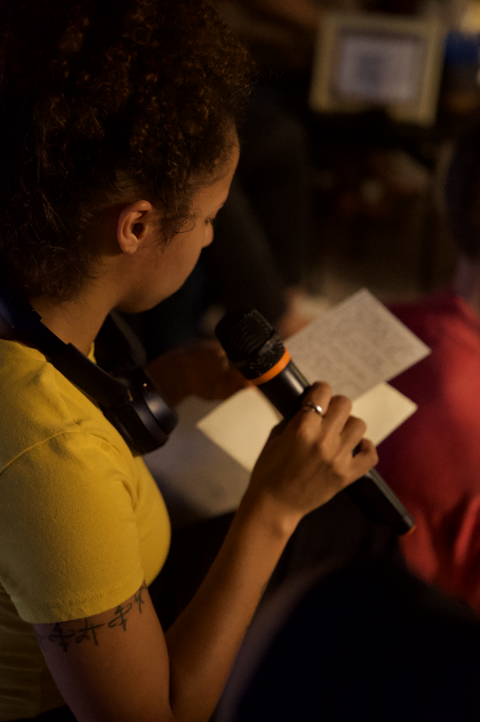 A woman with curly hair, wearing a yellow shirt and headphones around her neck, is reading from a piece of paper into a microphone.