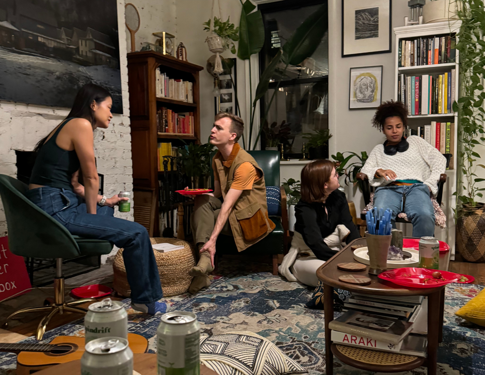 Four people in a cozy living room, engaged in conversation. The room has bookshelves, art, plants, and a small table with snacks and drinks. A guitar rests on the floor.