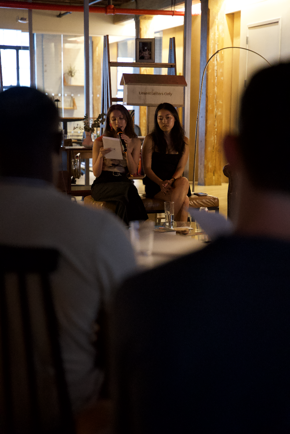 Two women sitting on a bench in front of an audience, one reading with a microphone, in a warmly lit room with a sign that says "Unsent Letters Only."
