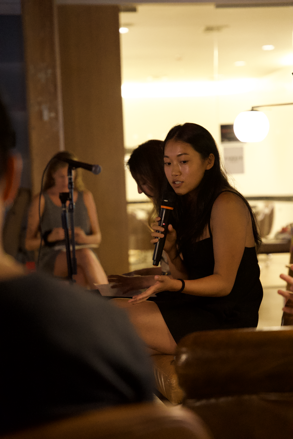 Woman in black dress speaking into a microphone during a presentation or discussion in a dimly lit room, with a seated woman in the background taking notes.