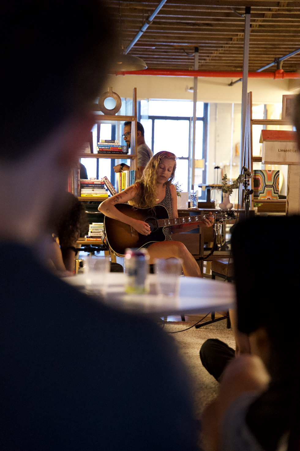 A musician playing acoustic guitar in a cozy indoor space, surrounded by bookshelves and an audience. The focus is on the performer, with blurred foreground elements and a warm atmosphere.
