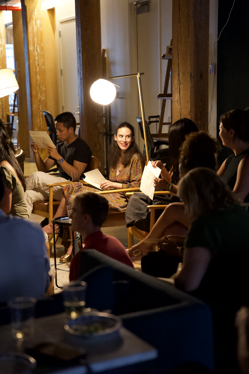 A group of people sitting on chairs in a cozy, warmly lit indoor setting, possibly a cafe or small theater, with some holding papers and engaging in conversation. The environment features exposed wooden beams and a floor lamp providing soft lighting.