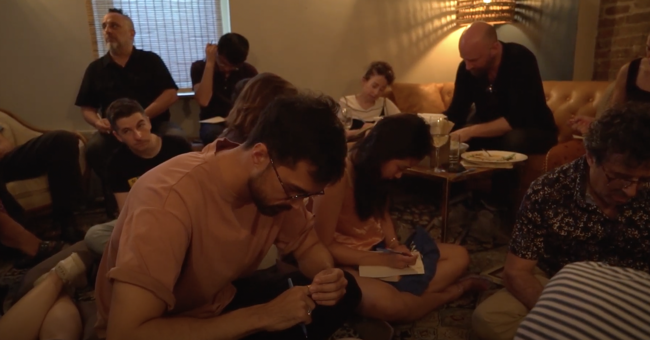 Group of people sitting on the floor and on couches, some writing or taking notes, in a cozy, dimly lit room with warm lighting and brick walls.