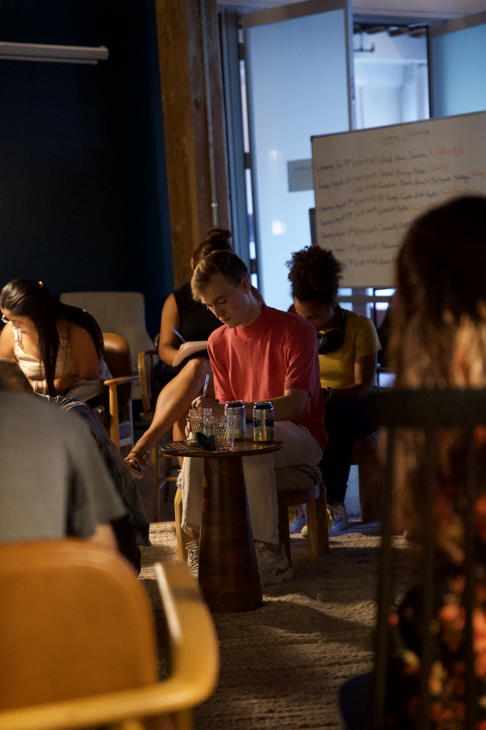 A group of people sitting in a dimly lit room, some taking notes, with a whiteboard in the background, possibly attending a workshop or seminar.