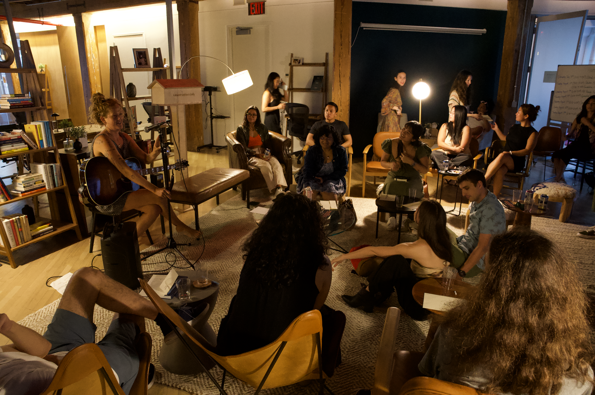 Group of people sitting and listening to a musician playing guitar in a cozy indoor setting.