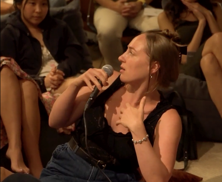 A person with a microphone speaking at a gathering, surrounded by seated audience members.