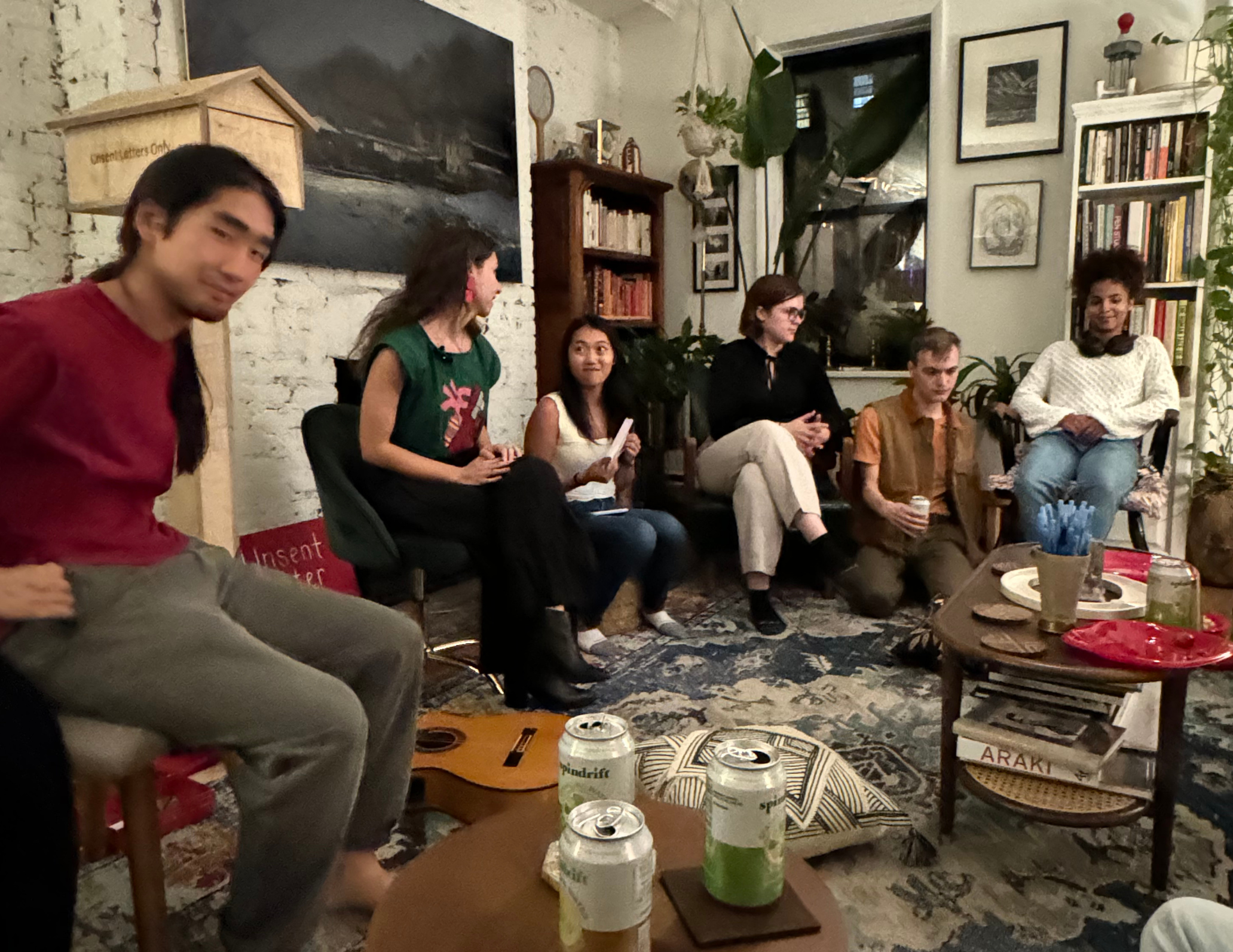 A group of people sitting in a cozy living room having a discussion. The room has a bohemian style with plants, bookshelves, and art on the walls. A guitar is on the floor, and there are drinks and snacks on a coffee table.