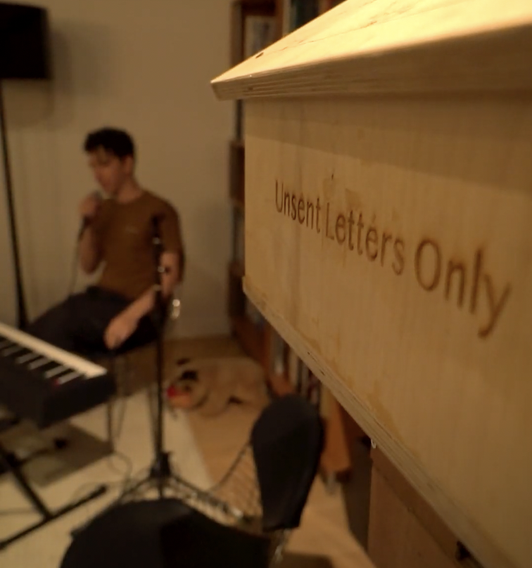 Close-up of a wooden box with 'Unsent Letters Only' inscribed, with a blurred background showing a person sitting near a keyboard.