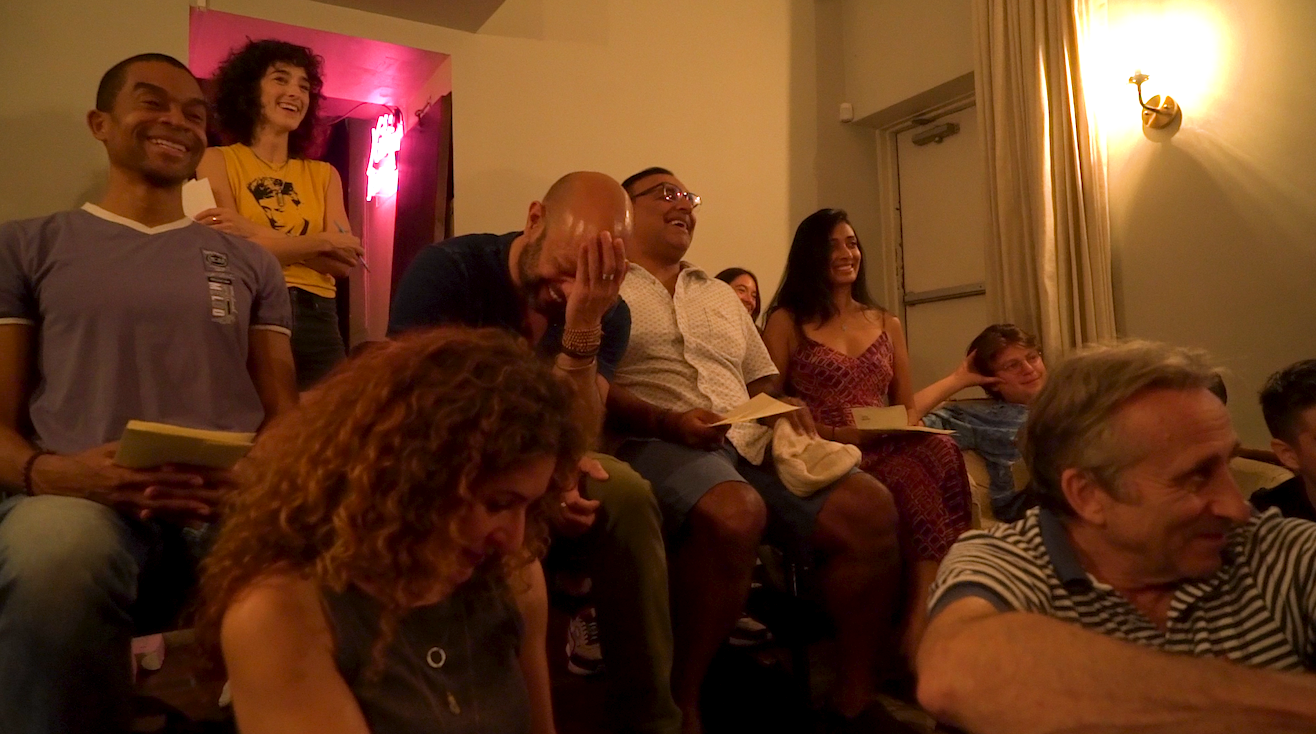 Group of people laughing and smiling in a cozy, dimly lit room, some holding papers and sitting on chairs.