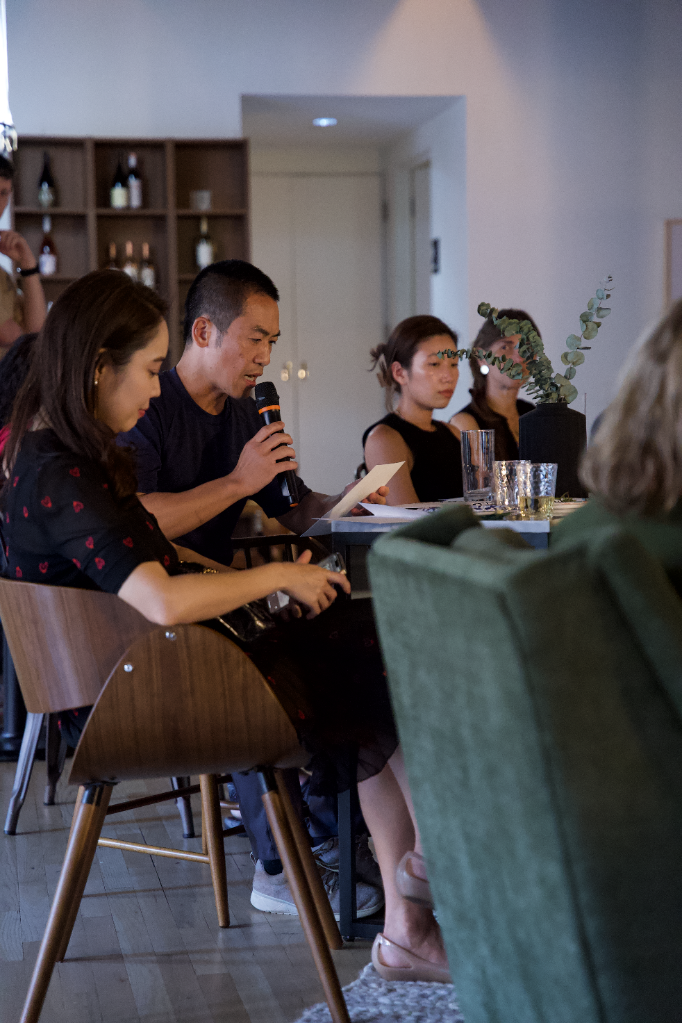 A group of people seated around a table in a room with dim lighting. One person is speaking into a microphone while reading from a piece of paper. Others are listening attentively. The setting includes chairs, a table with glasses, and shelves with b