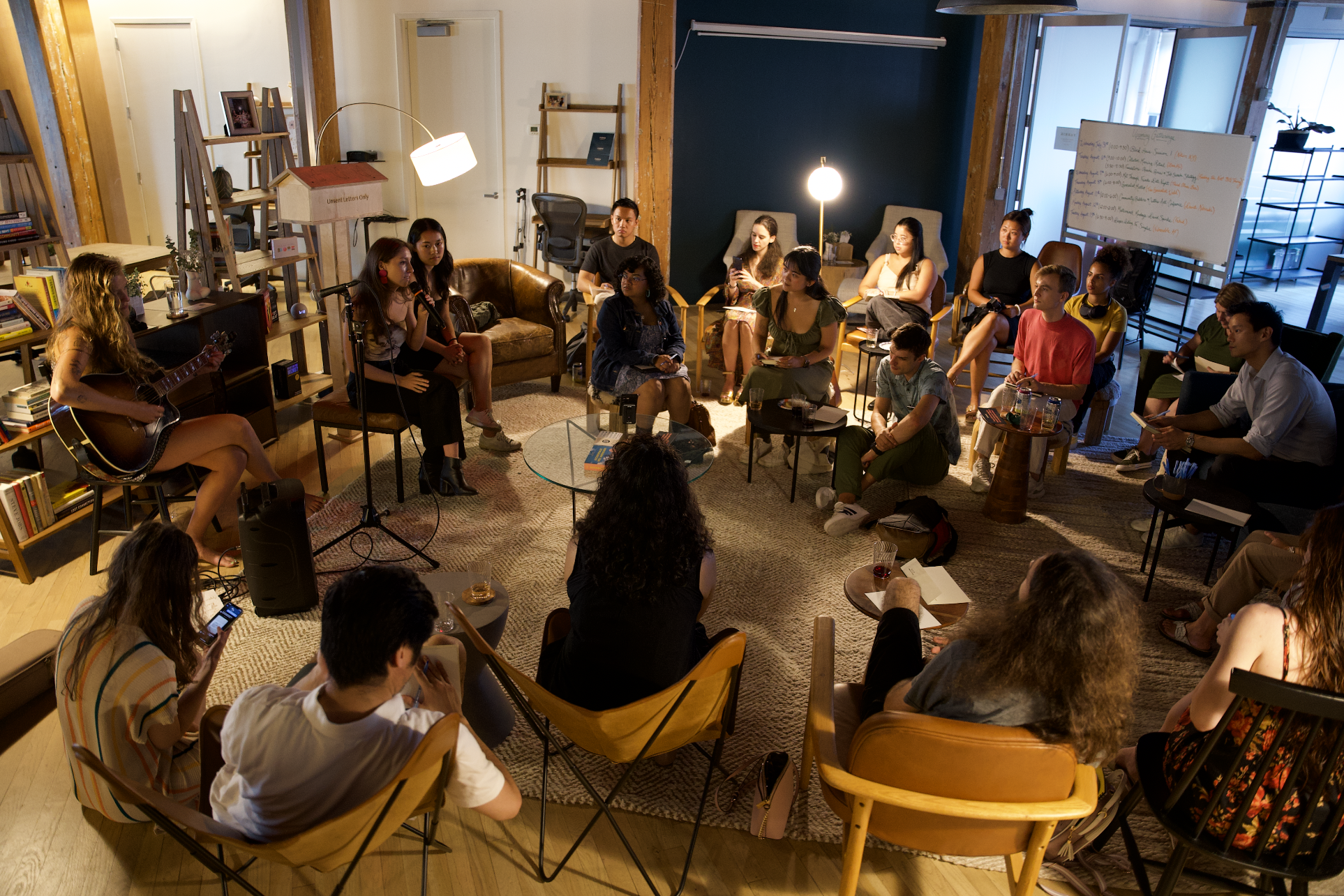 A group of young people gathered in a cozy living room for a small performance, with one person playing guitar and two others singing into microphones. The audience is seated in a semi-circle, some taking notes or using phones, with coffee tables hol