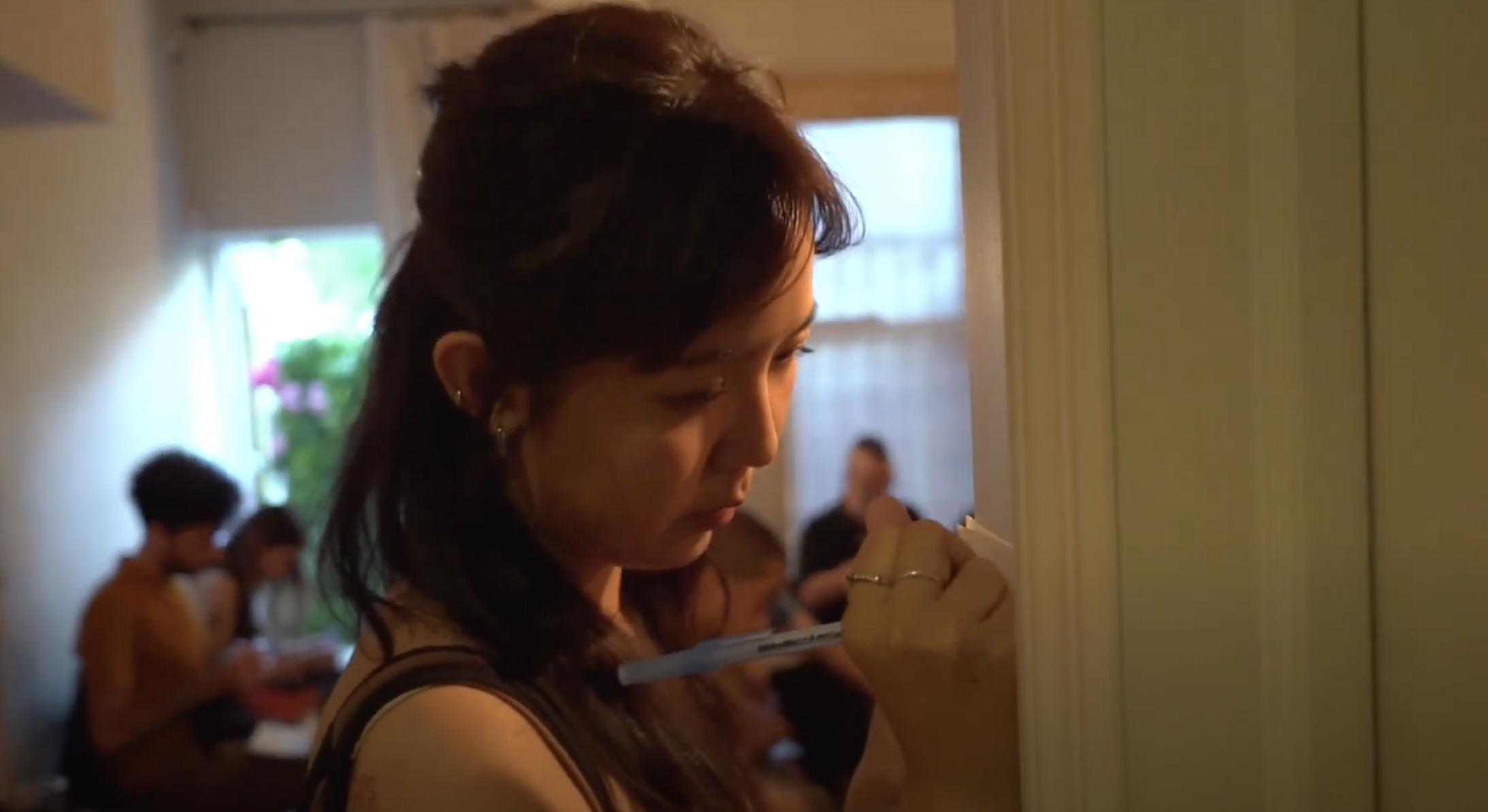 A woman with dark hair is writing on a wall or door with a marker inside a room, with several people seated in the background, some reading or writing.