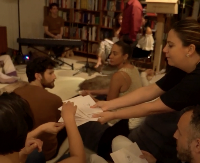 Group of people sitting in a cozy room, exchanging papers, with a keyboard in the background and bookshelves lining the walls.
