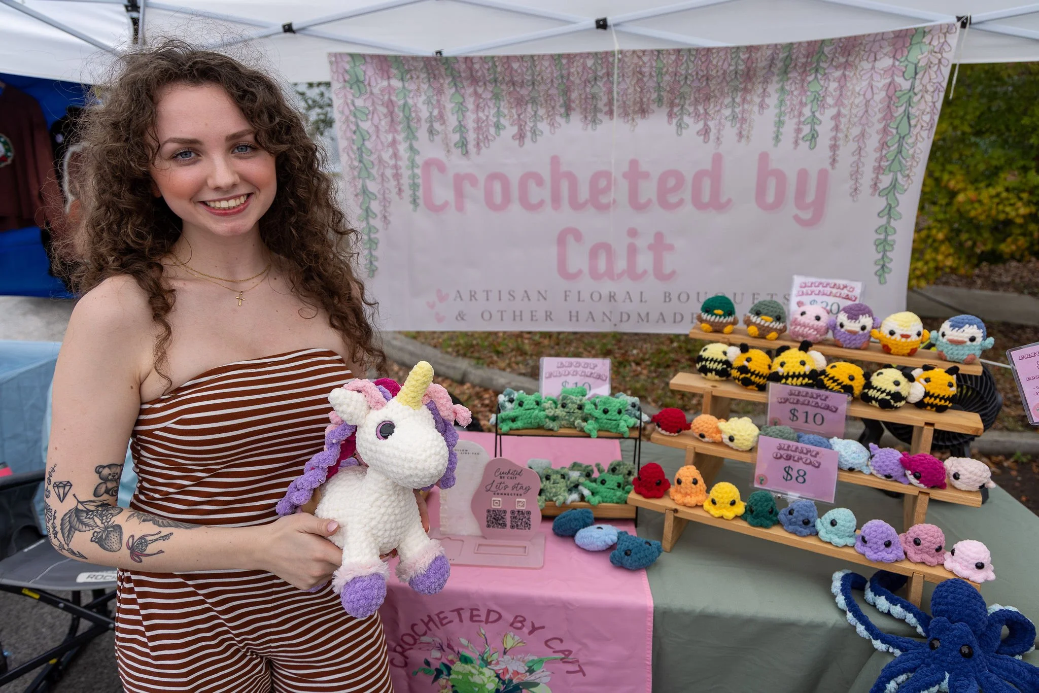 Woman with curly hair and tattoos on her arm smiling and holding a crocheted unicorn toy at a craft booth. The booth displays various handmade crocheted items, including small animal plushies and accessories, with a sign saying 'Crocheted by Cait'.