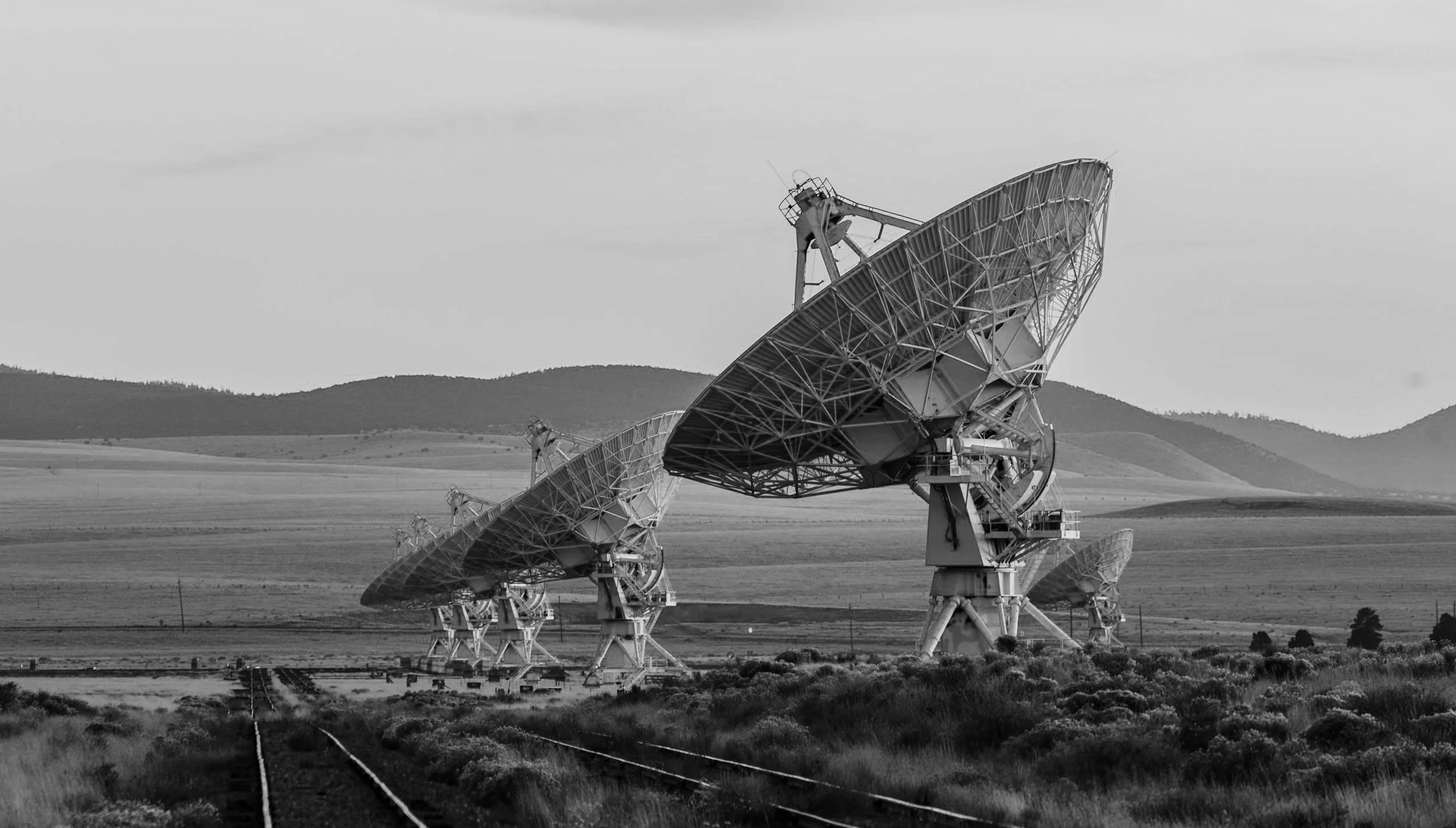 Three large satellite dishes in a field with rolling hills in the background, black and white.