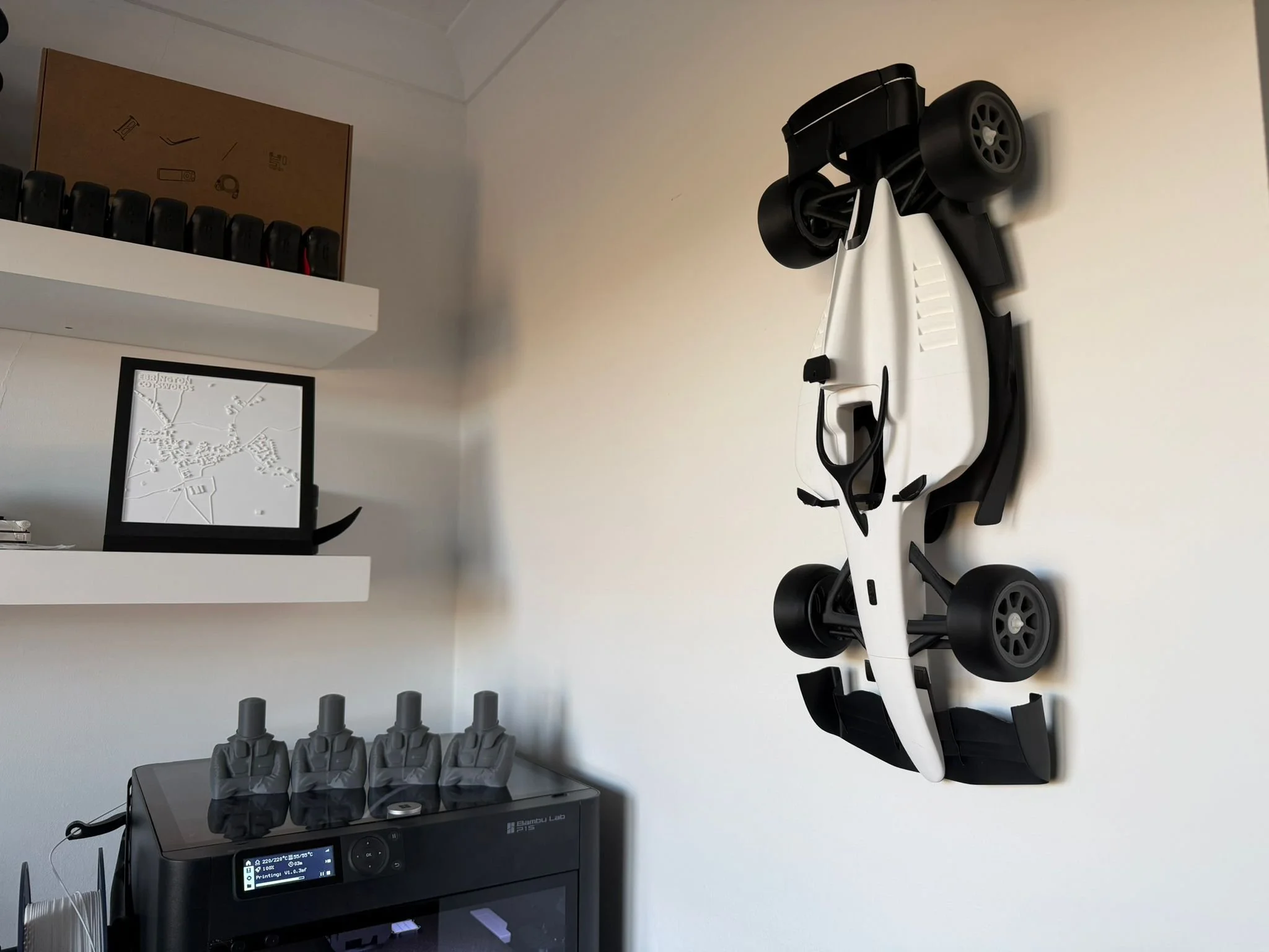 A white 3D-printed F2 inspired racecar model. hanging from a wall next to a 3D-printer