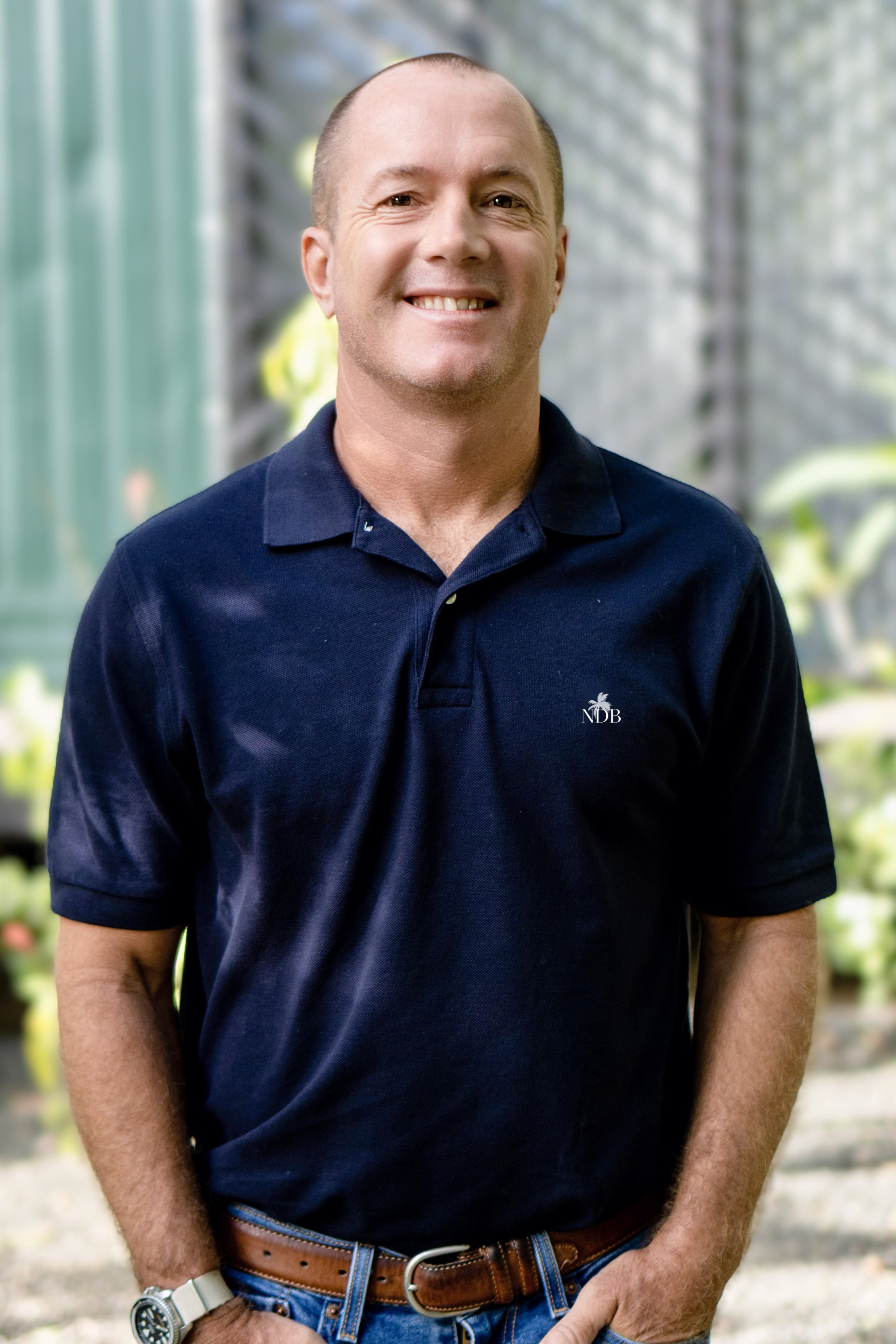 A middle-aged man with short hair smiling, wearing a navy blue polo shirt with a small logo, a leather belt, blue jeans, and a white wristwatch, standing outdoors with blurred greenery and a greenhouse in the background.