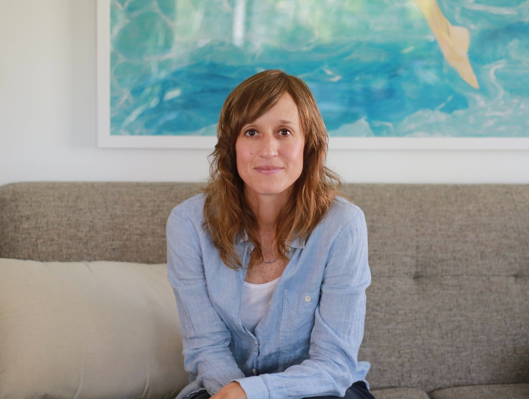 Woman with long hair sitting on a couch in front of a painting featuring blue and green tones.