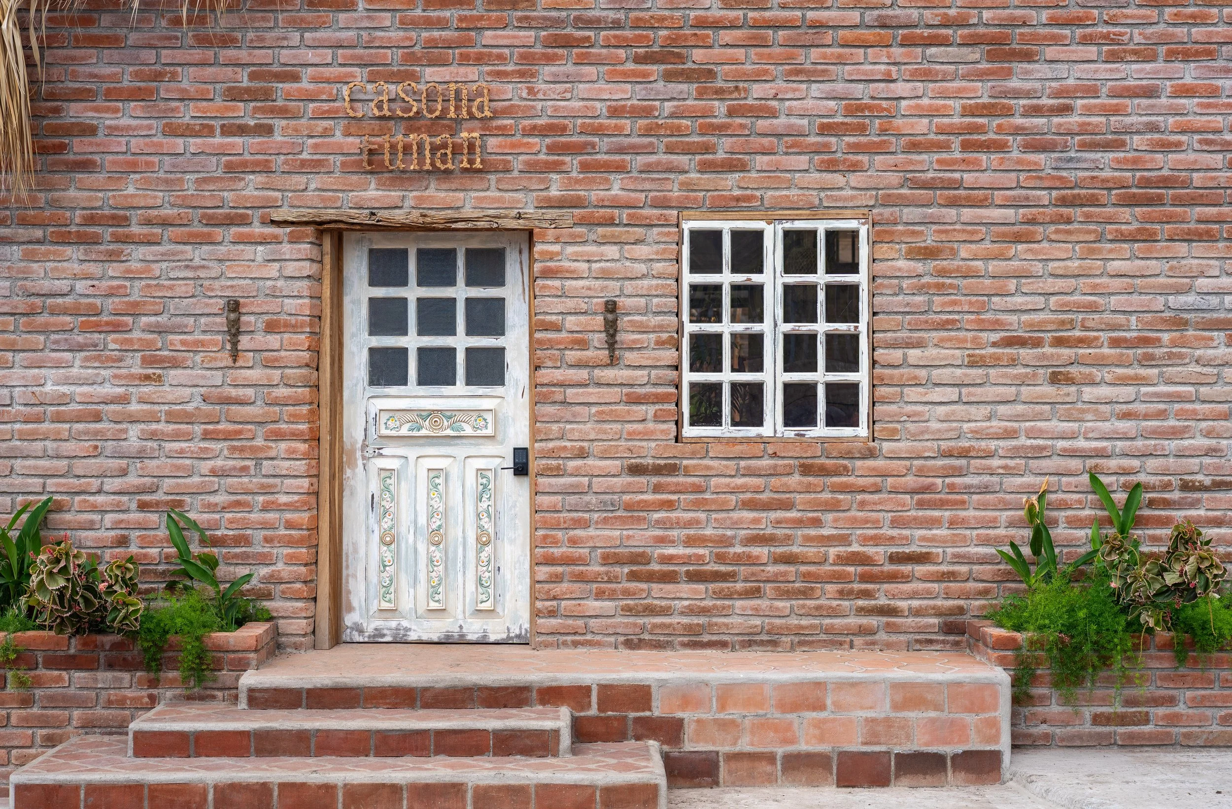 A brick house entrance with a wooden door featuring decorative painted details and a black electronic door lock. There are three steps leading up to the door, with green plants and flowers on both sides. To the right of the door is a window with whit