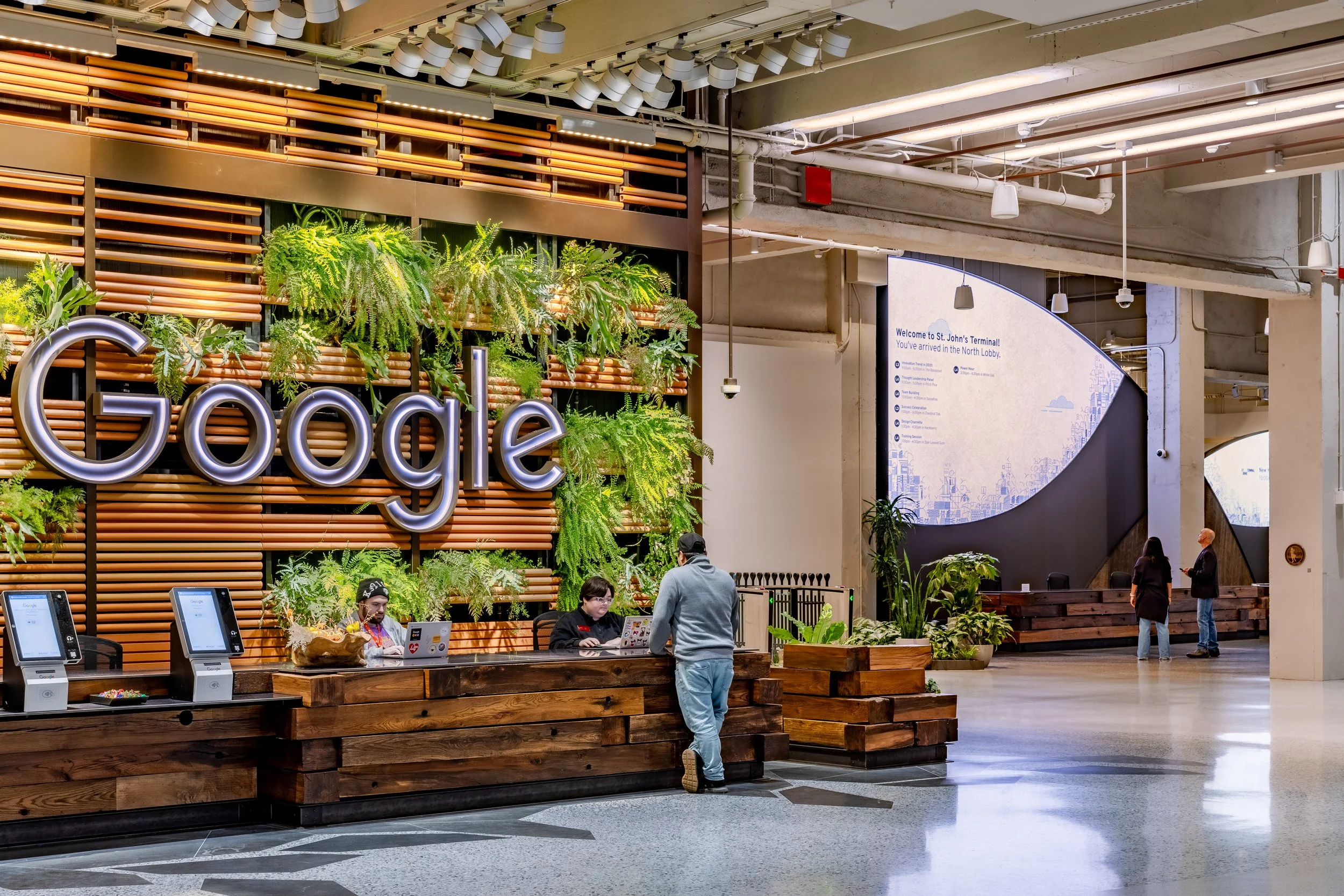 Indoor area at Google with a wooden reception desk, green plants, and people working or waiting, and a sign welcoming visitors to St. John's Terminal.