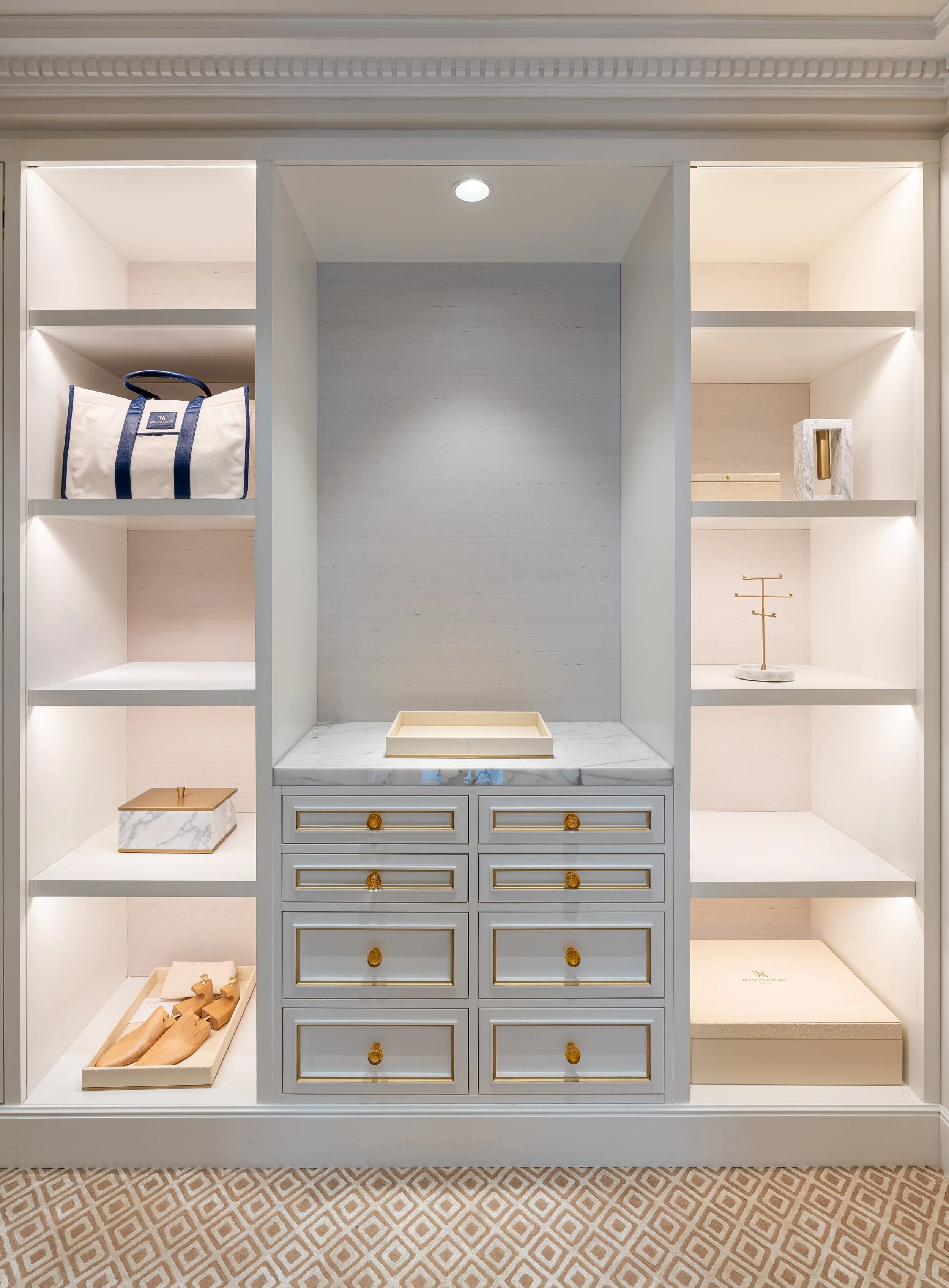 A built-in display shelf with multiple sections, featuring a white bag with blue handles, wooden objects, and decorative items on white shelves and drawers.