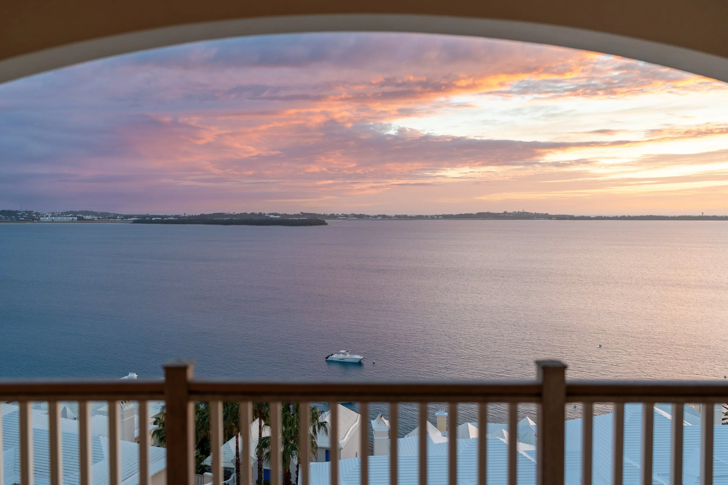 View of a sunset over a body of water, seen from a balcony with railings and white rooftops below.