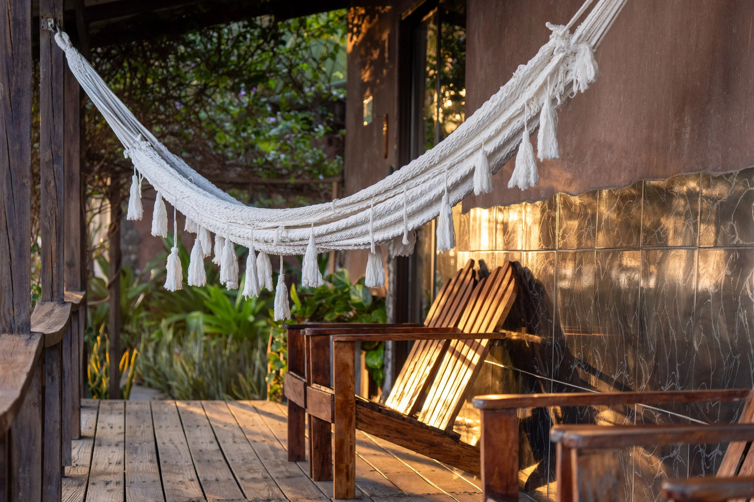 A cozy outdoor patio with a white hammock hanging between wooden posts, surrounded by greenery, and warm lighting illuminating the space.