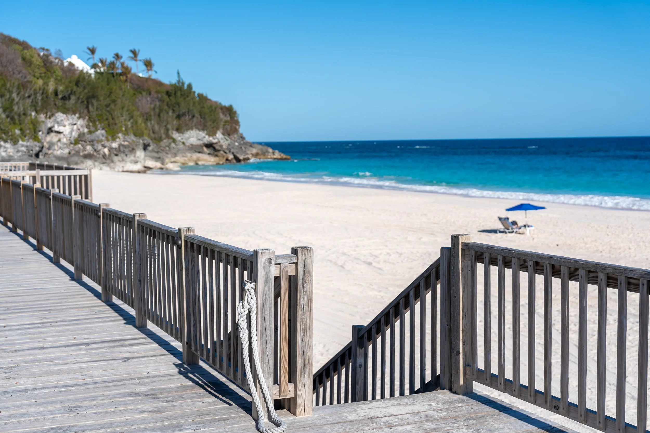A wooden boardwalk leading to a sandy beach with a view of the ocean, a single blue umbrella, and a lounge chair. There are rocky cliffs with trees in the background and a bright blue sky.