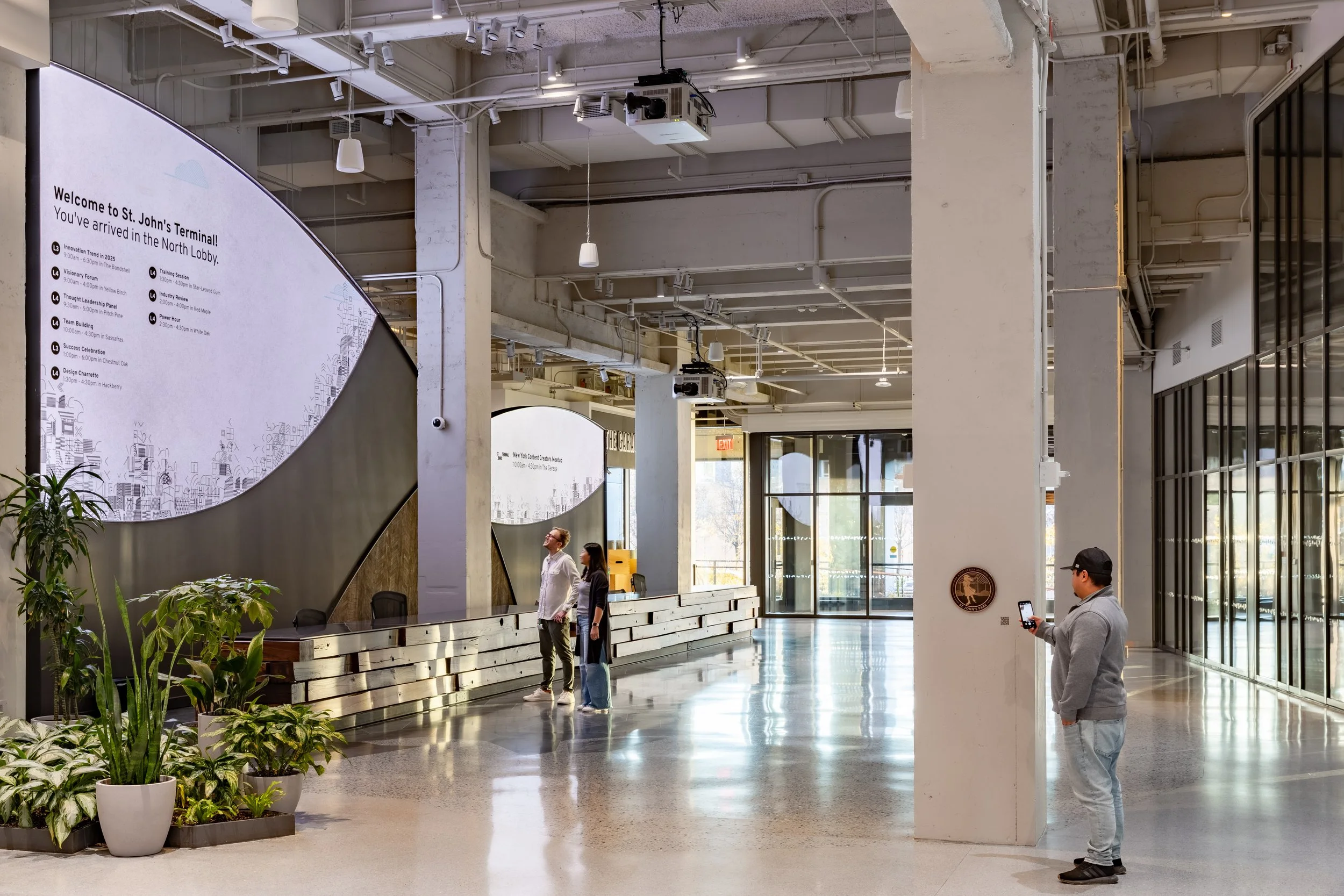 Interior of a modern transportation terminal with a large digital welcome sign, a check-in or information counter, and several people. There are plants in white pots, exposed ceiling pipes, and large windows letting in natural light.