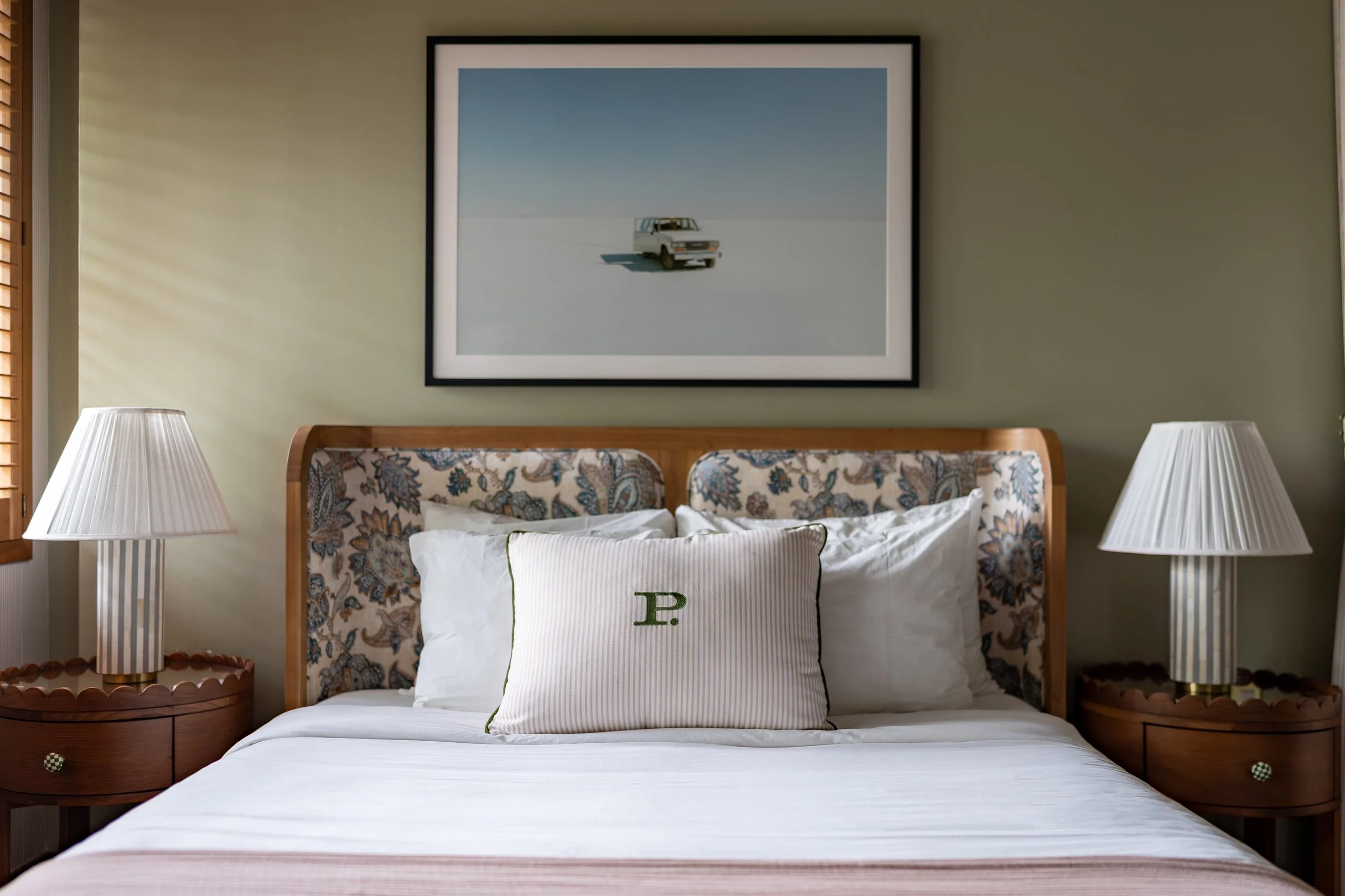 A neatly made bed with white sheets, a decorative pillow embroidered with the letter 'P', and a floral patterned headboard. Above the bed, there is framed artwork of a white vehicle in a minimalist landscape. On either side of the bed, there are matc