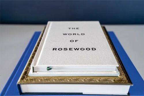 A book titled 'The World of Rosewood' resting on a decorative gold-colored frame on a blue surface.