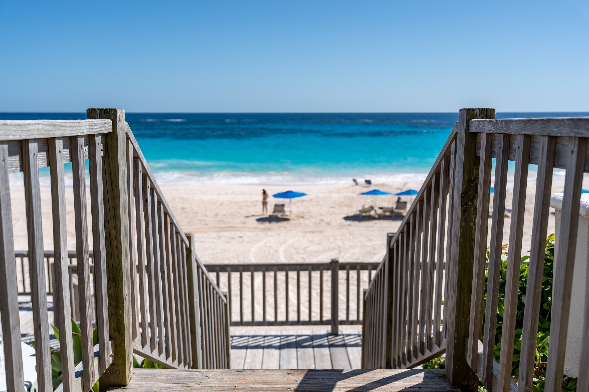 View of a beach from wooden stairs with railings, showing sandy shore, umbrellas, and people on the beach with the ocean and clear blue sky in the background.