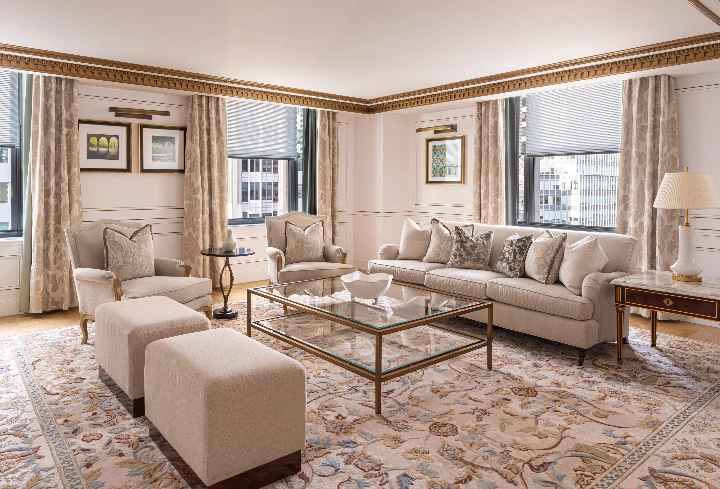 Elegant living room with cream-colored couches and armchairs, a glass coffee table, ornate patterned area rug, framed artwork on white walls, and large windows with patterned curtains, cityscape view outside.