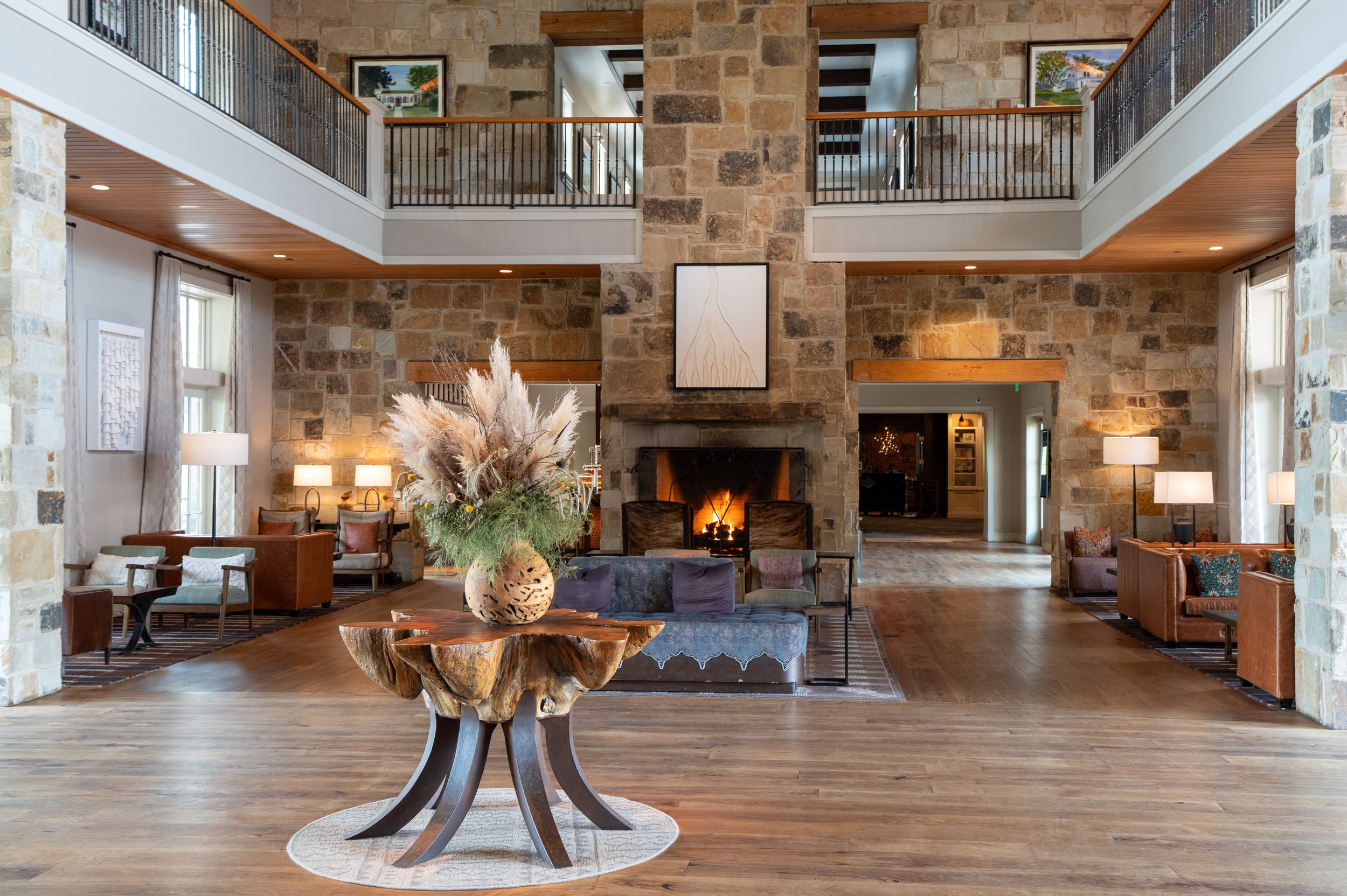 A spacious living room with a stone fireplace, wooden floors, and high ceilings. A large floral arrangement is on a wooden table in the center, and various modern seating options are arranged around it. There is a balcony with black metal railings on