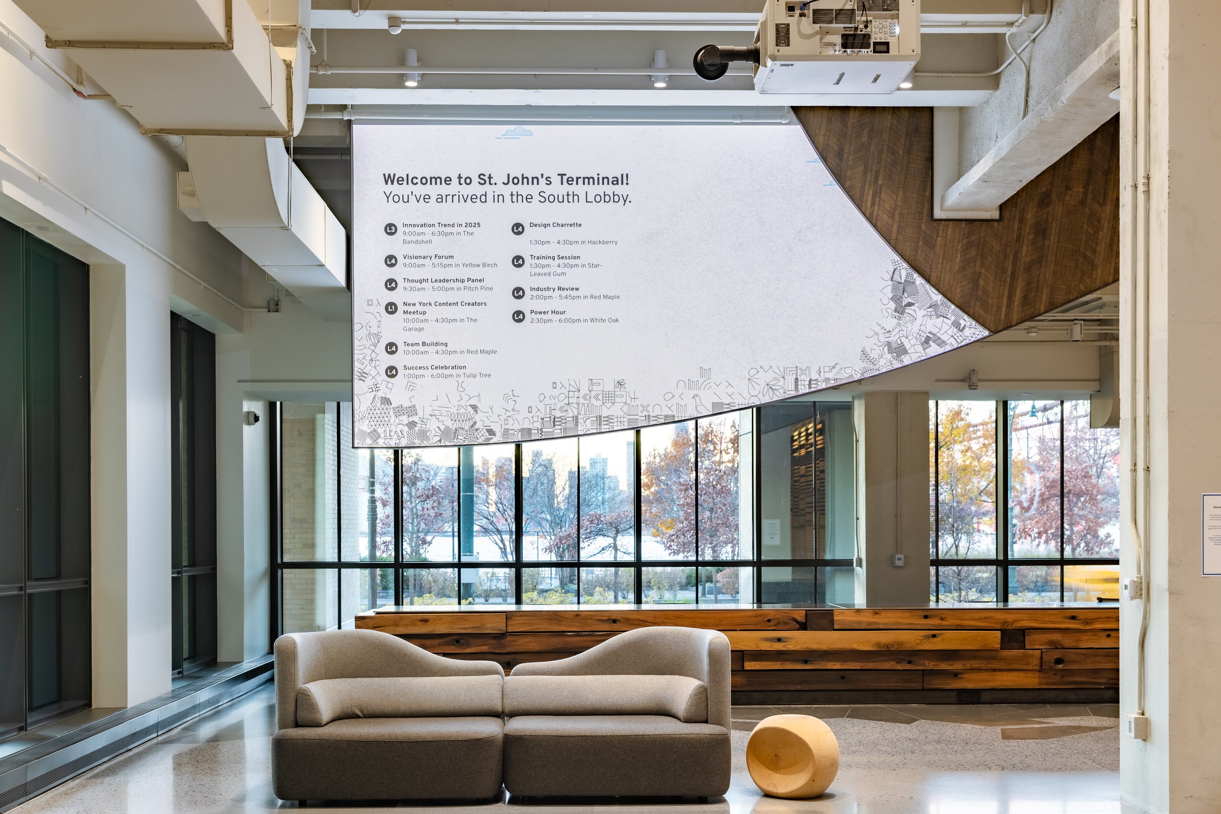Lobby area with large digital screen displaying a welcome message for St. John's Terminal, and a seating area with a beige sofa and a round wooden decorative piece in front of it. Large windows reveal trees with autumn foliage outside.