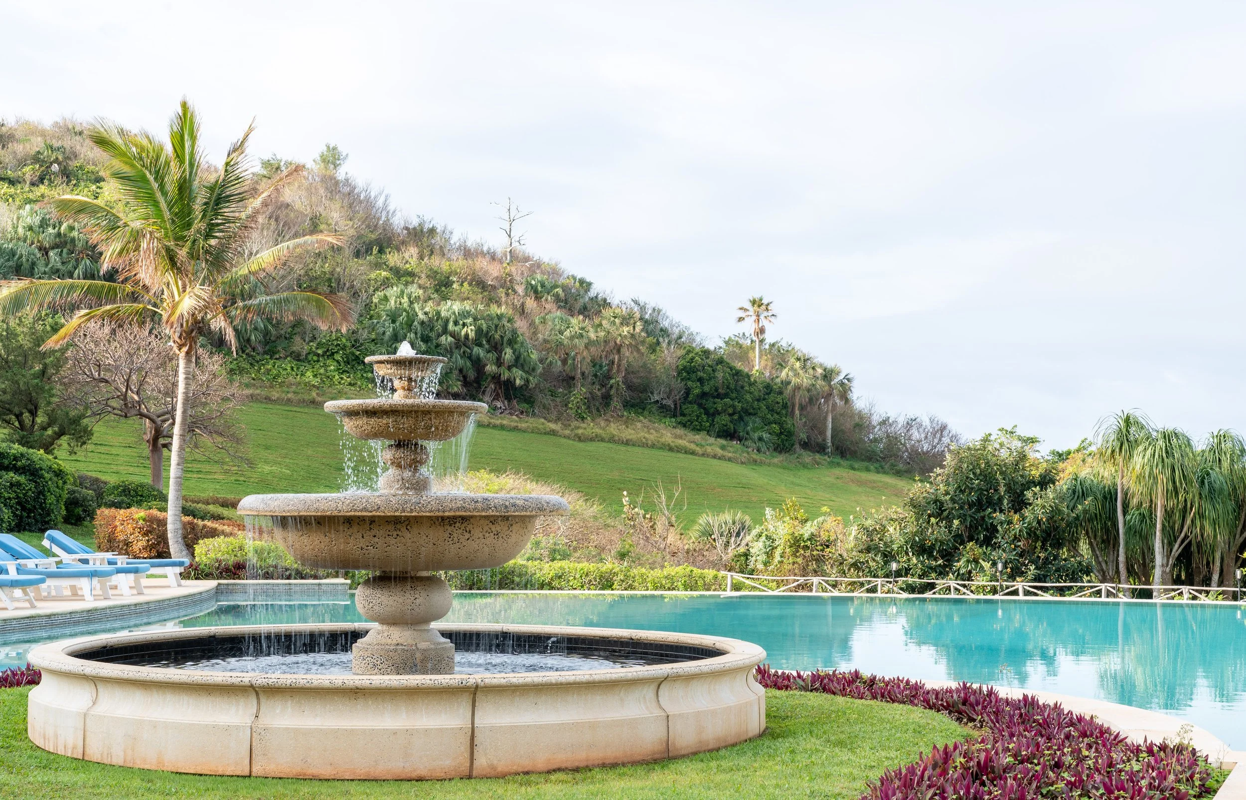 A decorative stone fountain with water cascading from the top tier, surrounded by lush green grass, tropical trees including palm trees, and colorful flower beds, with a hilly landscape in the background under a partly cloudy sky.