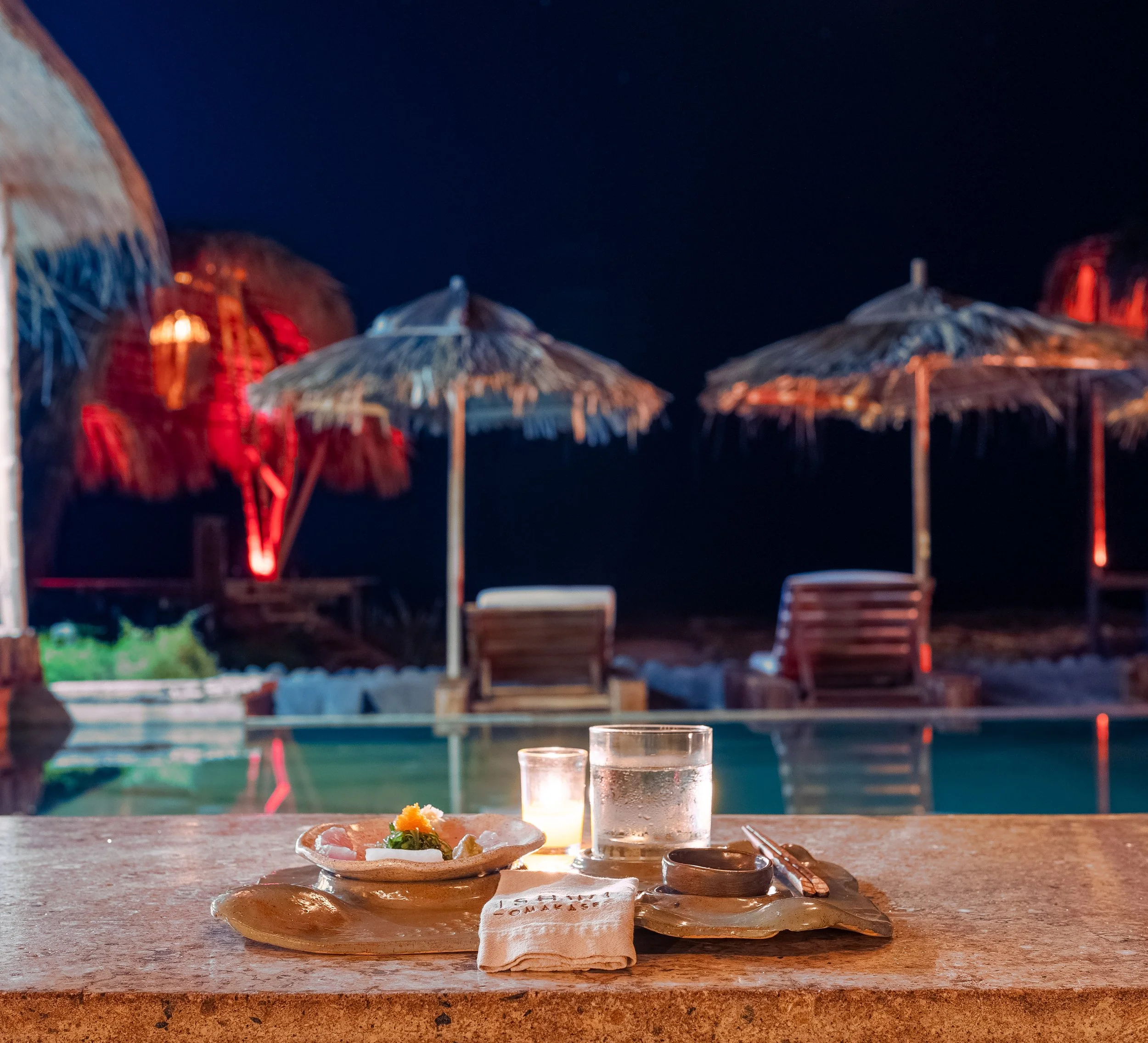 A table with a small plate of food, a glass of water, and a lit candle, set outdoors at night with thatched umbrellas and a dark sky in the background.