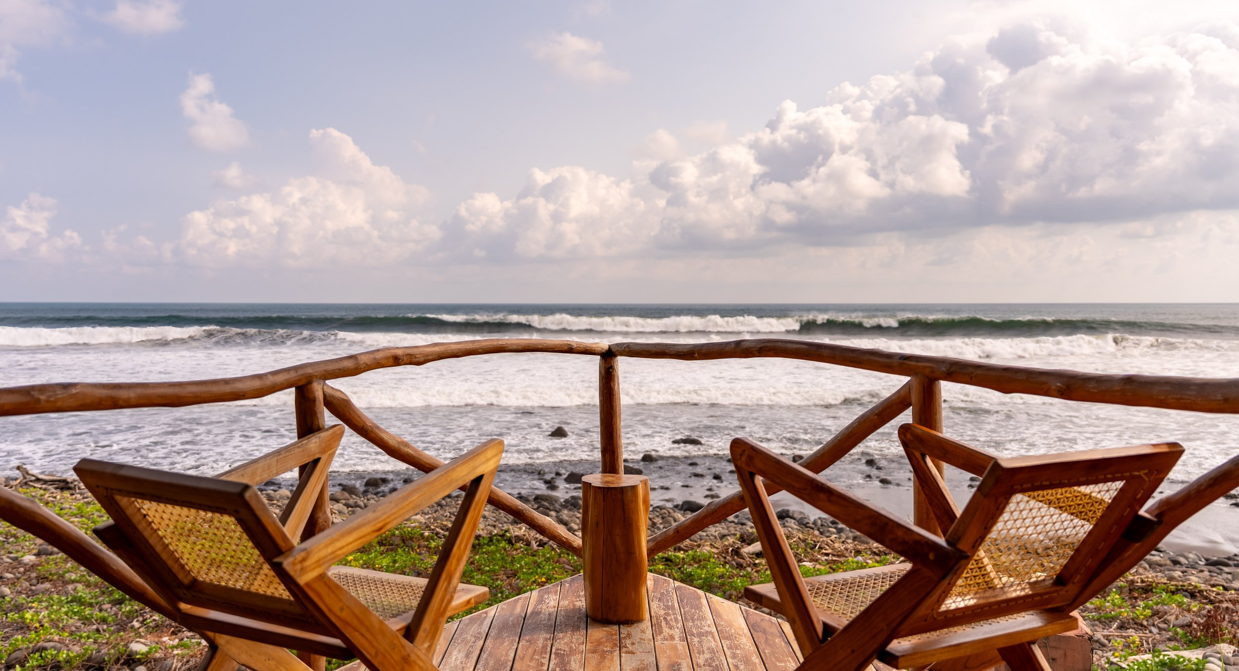 Beach scene with wooden chairs on a deck facing the ocean, with waves and a partly cloudy sky