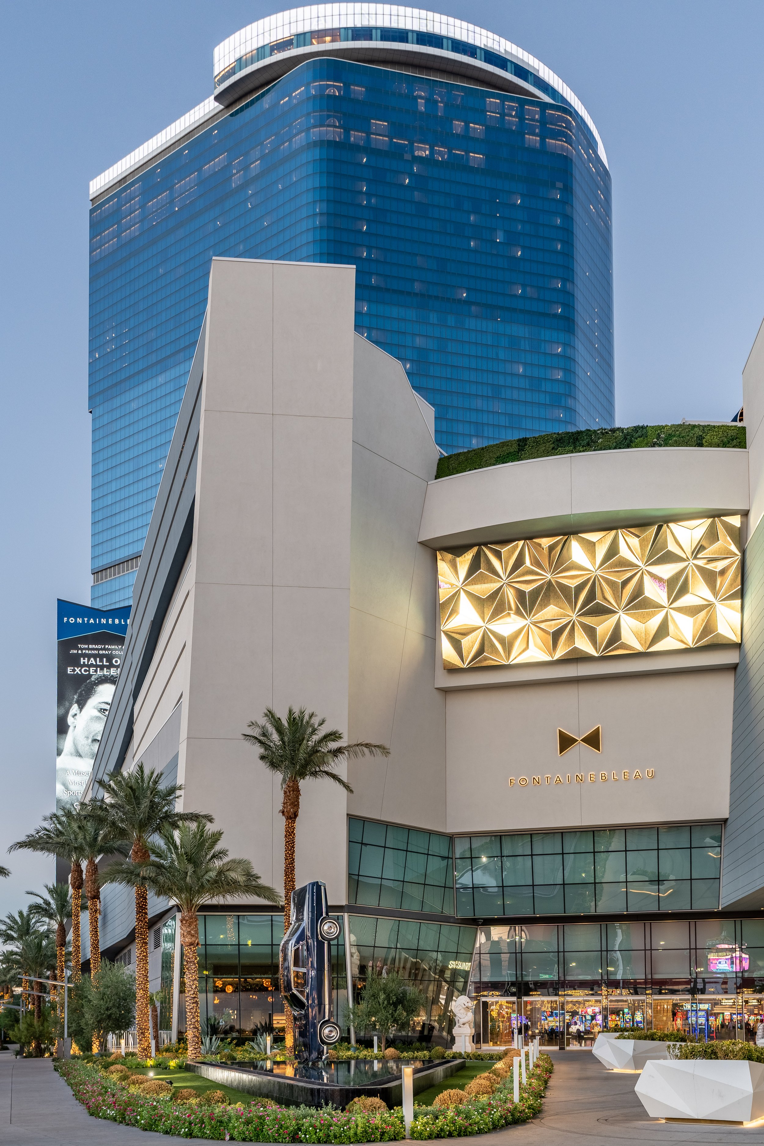 Exterior view of Fontainebleau hotel in Las Vegas, featuring a modern building with a blue glass facade, palm trees with festive lights, a gold geometric art piece, and a sculpture of a vintage car above a fountain. The hotel logo is visible below a 