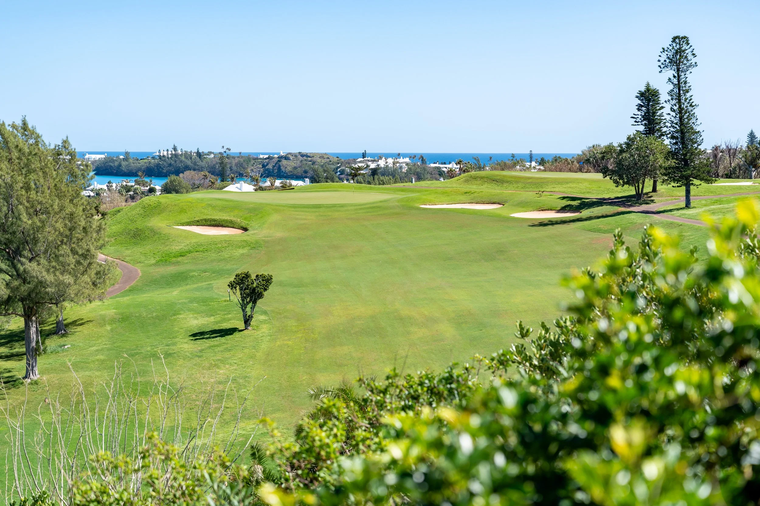 A scenic view of a golf course with green fairways, sand bunkers, and trees, overlooking a body of water under a clear blue sky.