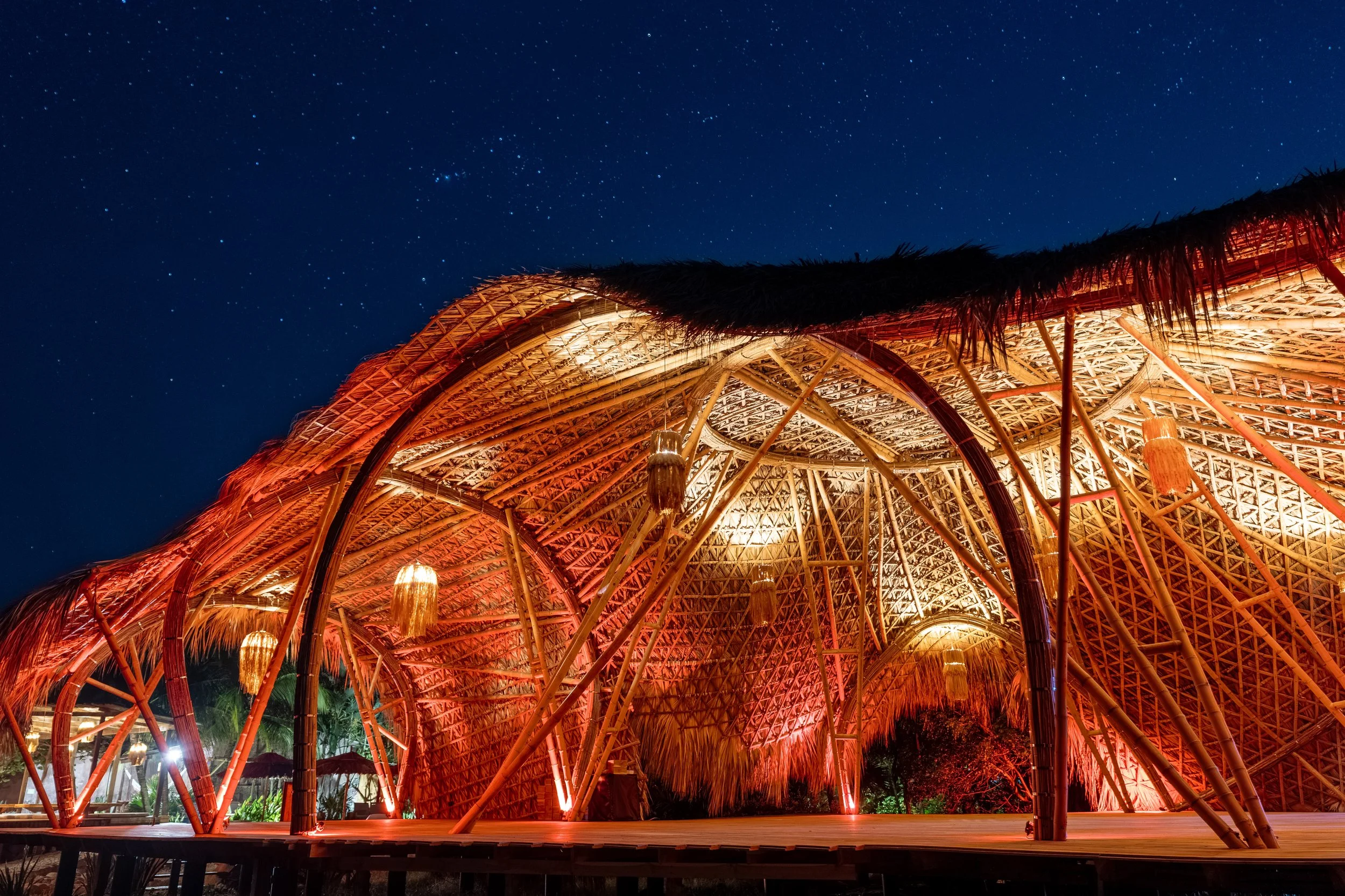 Nighttime view of a thatched-roof hut made of bamboo, illuminated from inside, with a starry sky overhead.