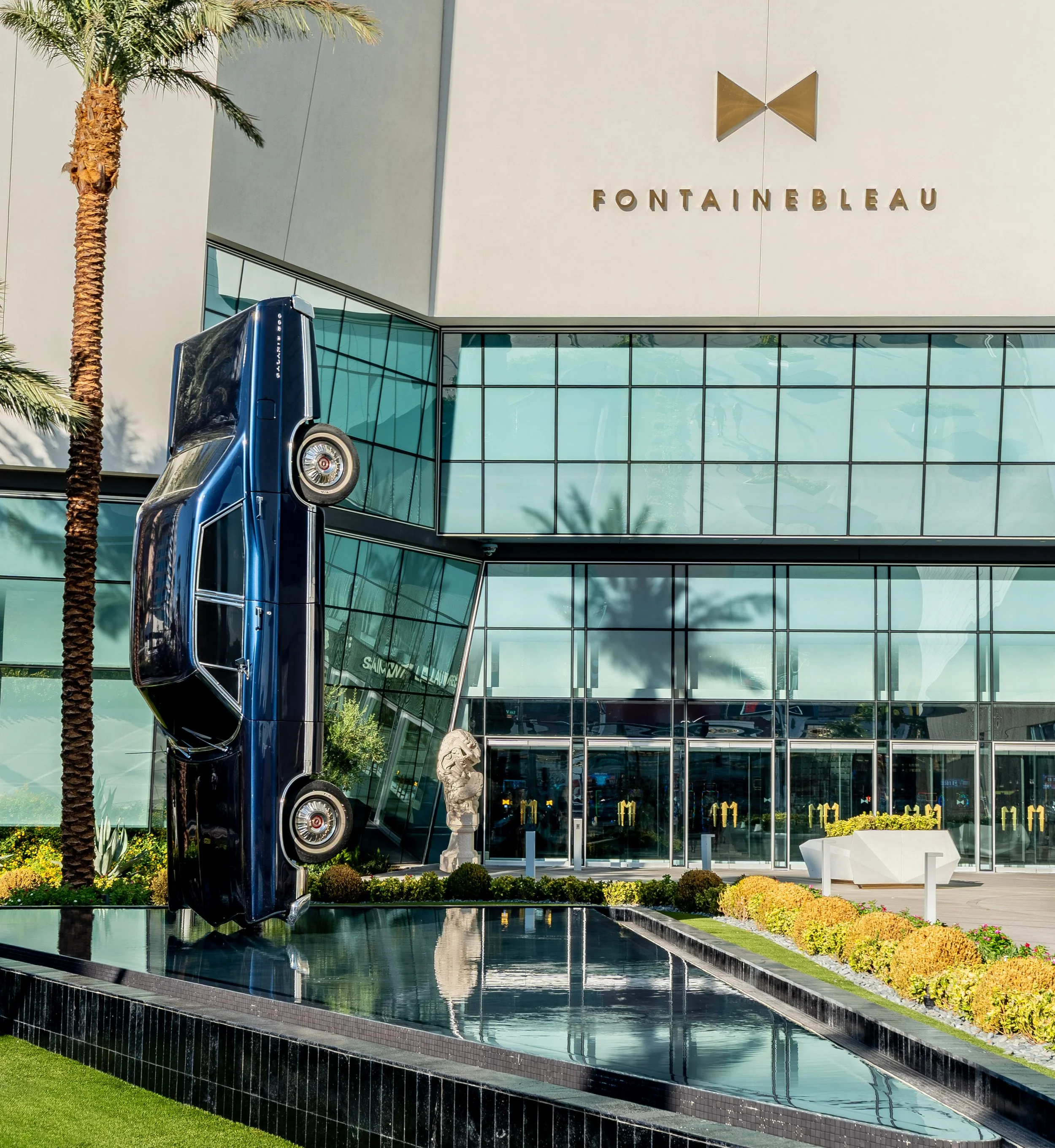 Modern building with glass windows and a sign that reads 'Fontainebleau'. A blue classic car is displayed outside, with a small pool or reflecting surface in front. There are landscaped bushes and flowers, a palm tree on the left, a stone lion statue