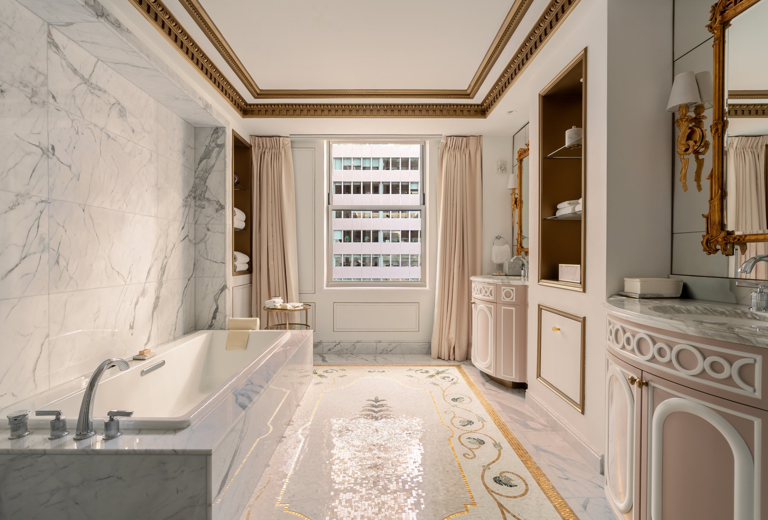 A luxurious bathroom with marble walls and floors, cream-colored curtains, a large window, gold-accented mirrors, and a bathtub.