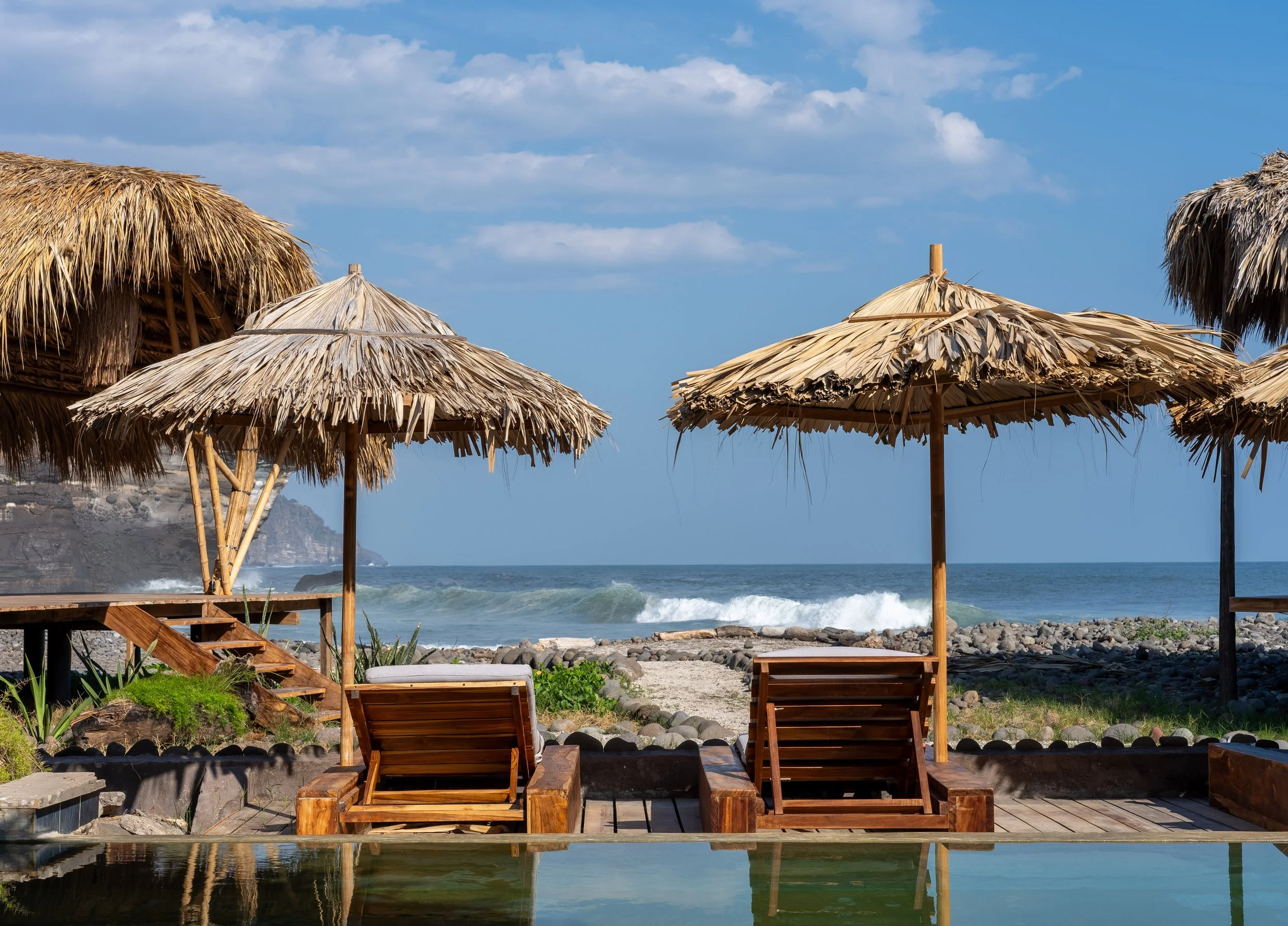 Beach scene with straw umbrellas, wooden lounge chairs, rocky shoreline, ocean waves, and cliffs in the background under a partly cloudy sky.